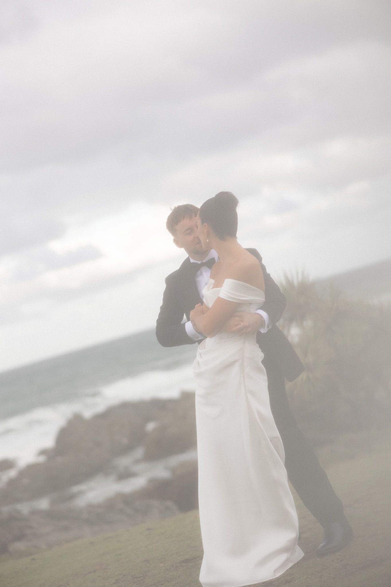 Bride and groom embrace romantically on a coastal clifftop, wearing wedding attire with ocean waves in the misty background.