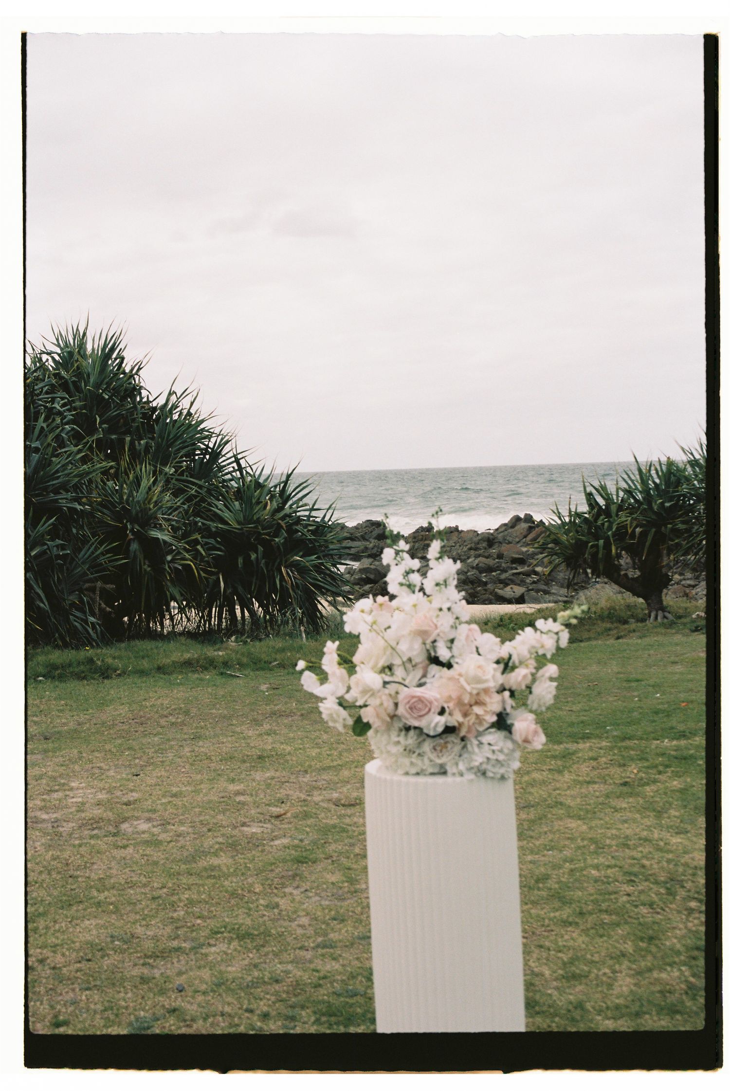 Elegant white floral arrangement on pedestal at outdoor beach wedding ceremony.