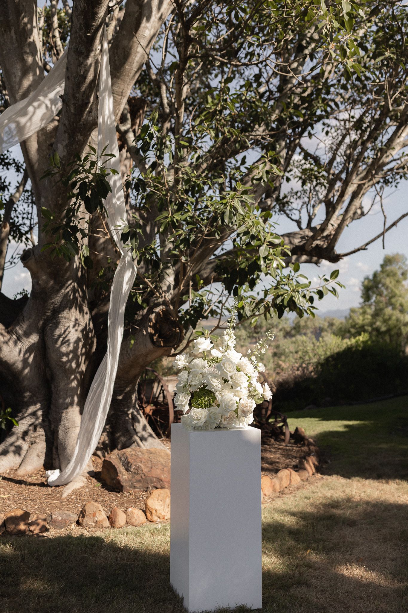 White floral arrangement on a pedestal beneath a large tree at an outdoor wedding ceremony.
