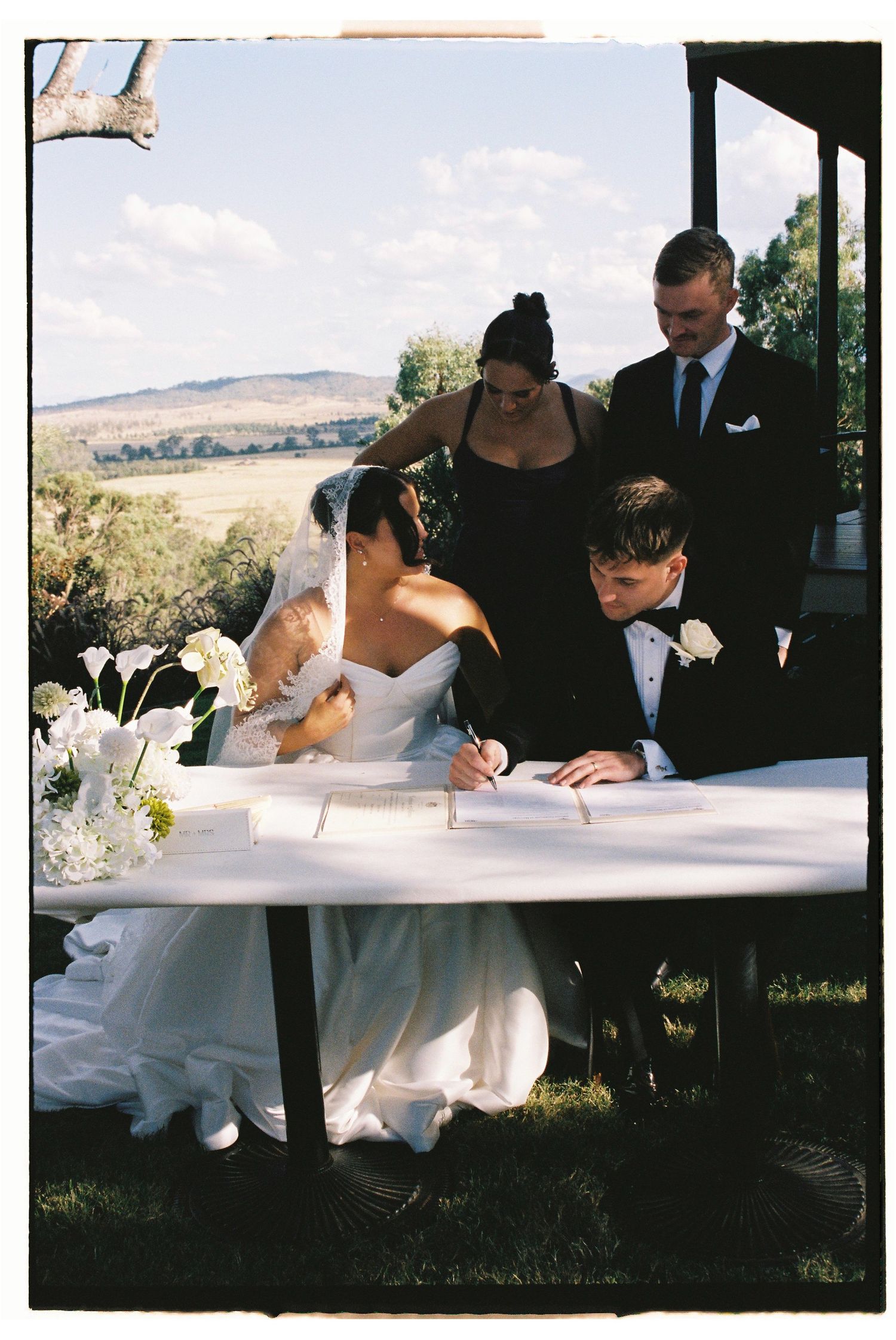 Bride and groom sign wedding register at outdoor table with witnesses standing behind.