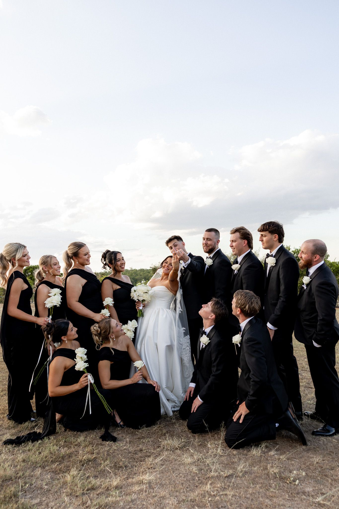 Wedding party in black attire surrounds bride and groom outdoors under cloudy sky at golden hour.