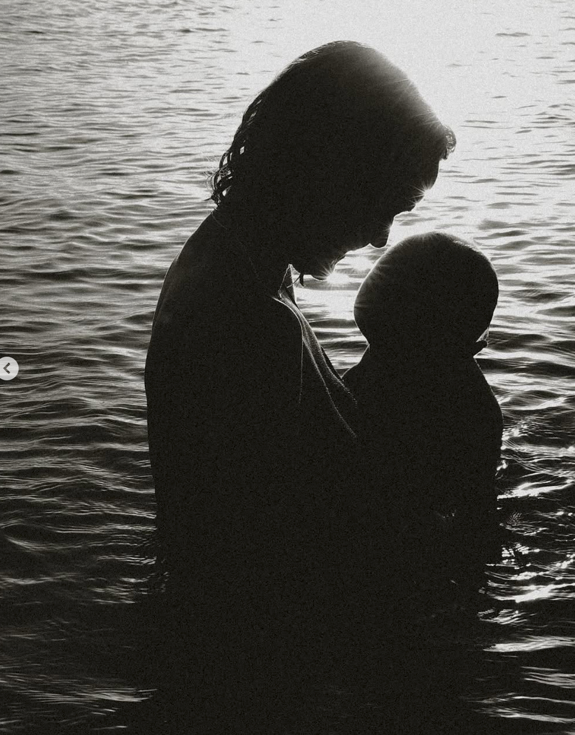 Black and white silhouette of a mother holding her baby near water, a tender moment captured in soft light.