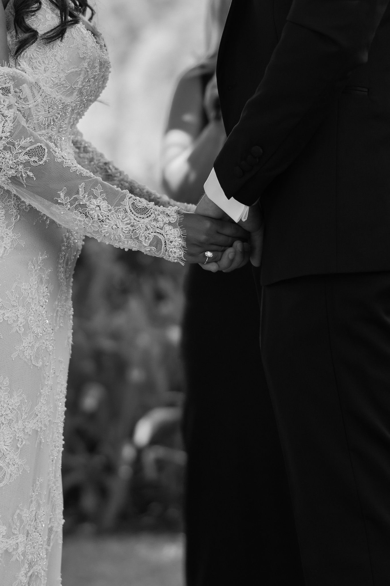Black and white photo of bride and groom holding hands during wedding ceremony, showcasing lace dress detail.