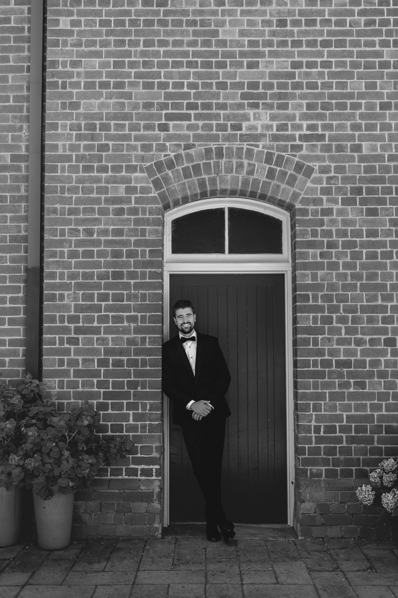 Black and white photo of a well-dressed man in a tuxedo standing in an arched brick doorway.