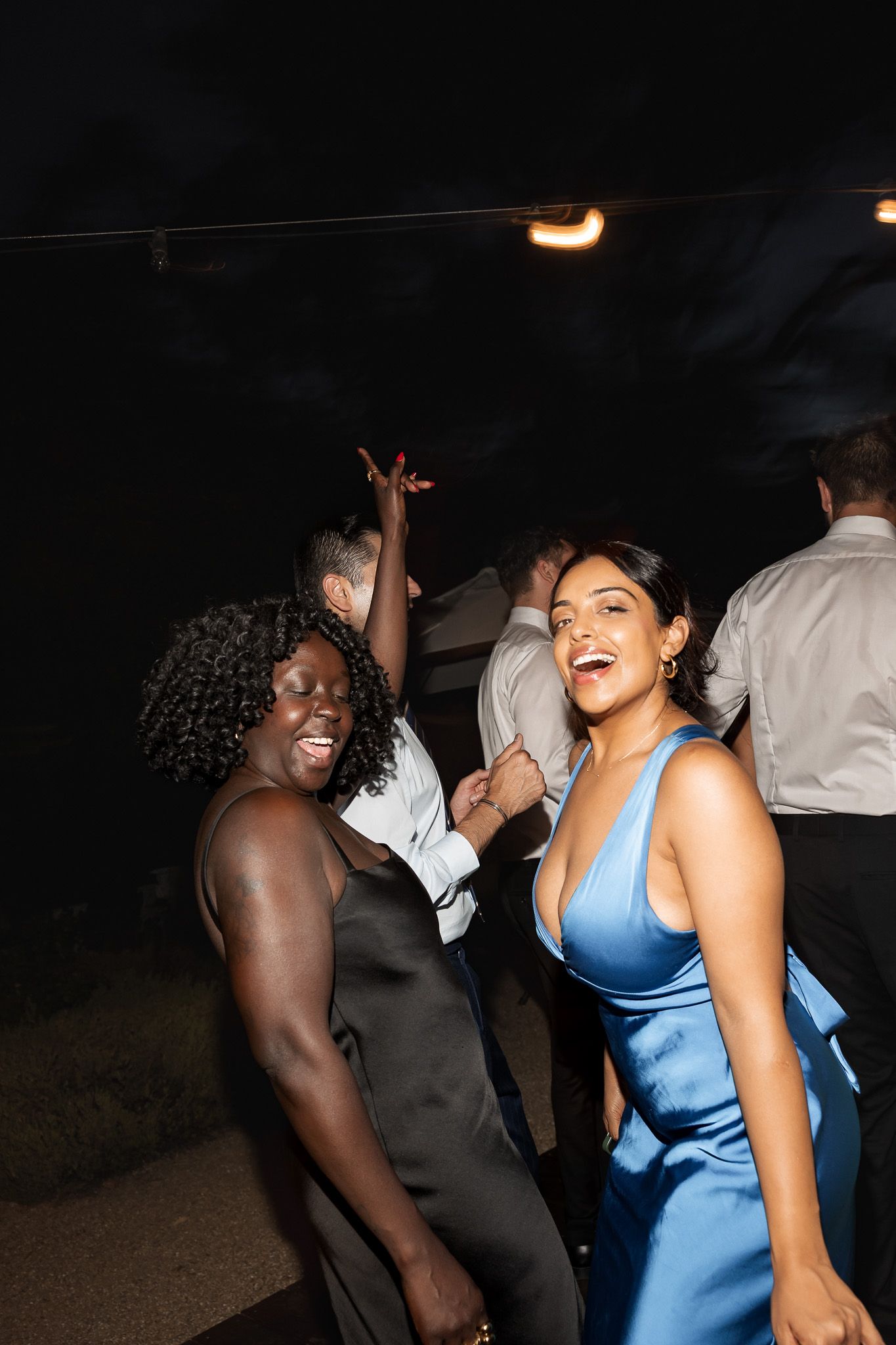 Two women in elegant dresses laughing and dancing at wedding reception.