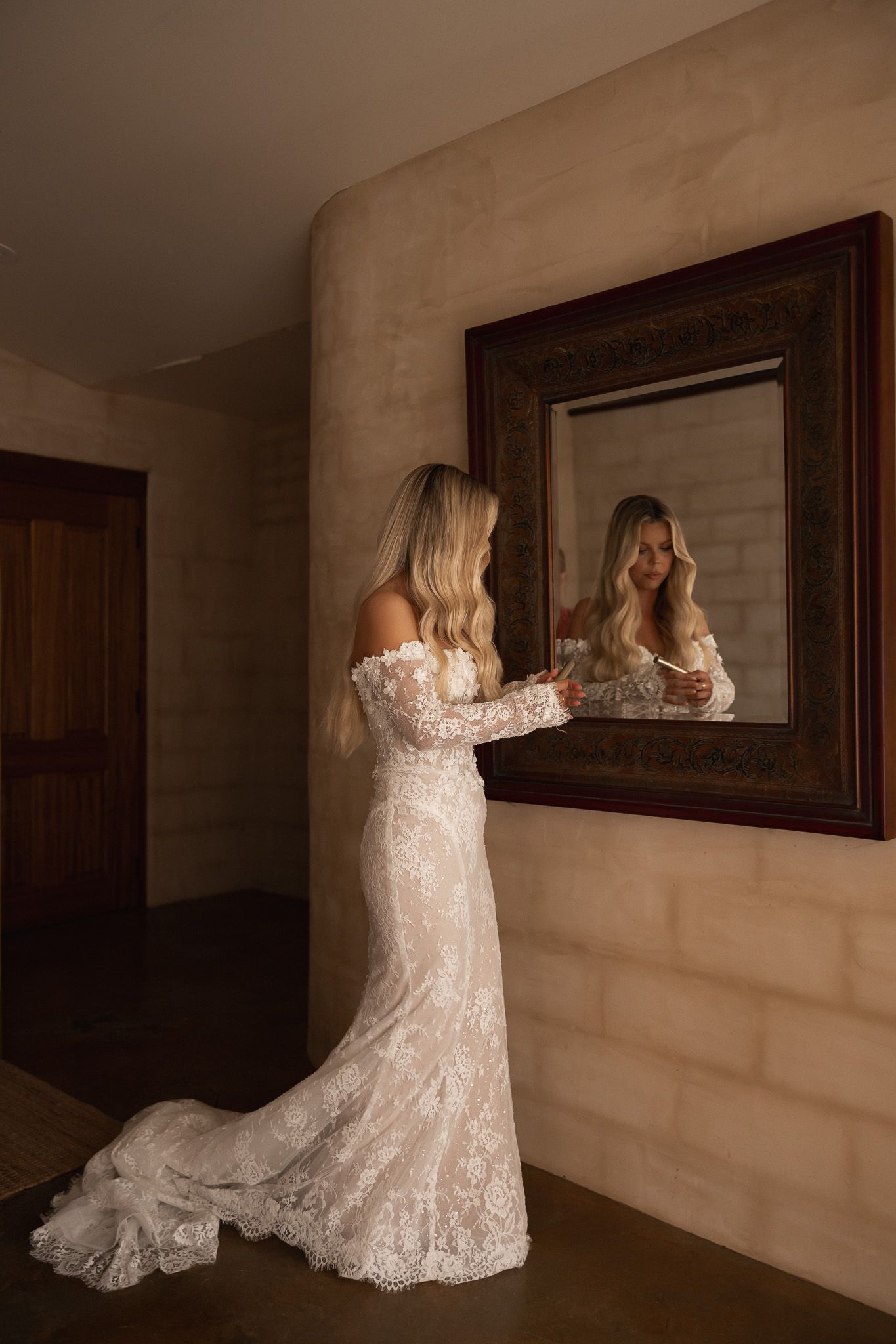 Bride in lace off-shoulder wedding gown admiring her reflection in ornate mirror in warmly lit room.