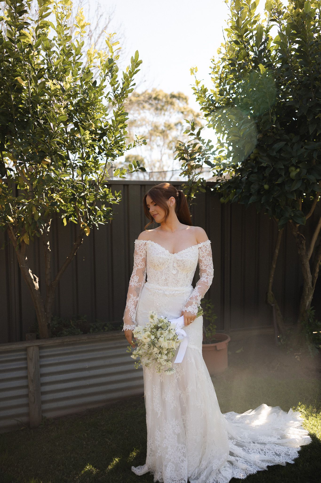 Bride in off-shoulder lace wedding gown holding white bouquet, standing between green trees in sunlit garden.