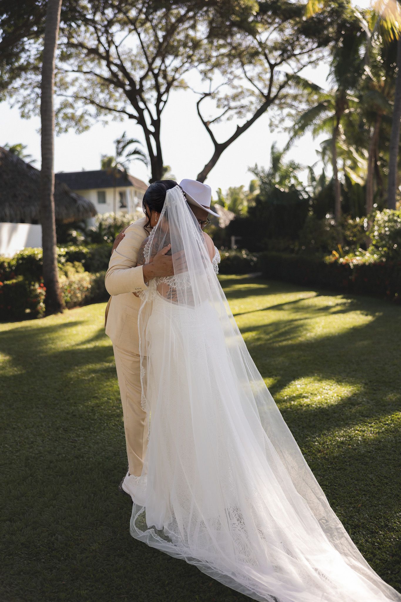 Bride and groom sharing romantic embrace on lush green lawn with tropical trees and thatched roof.
