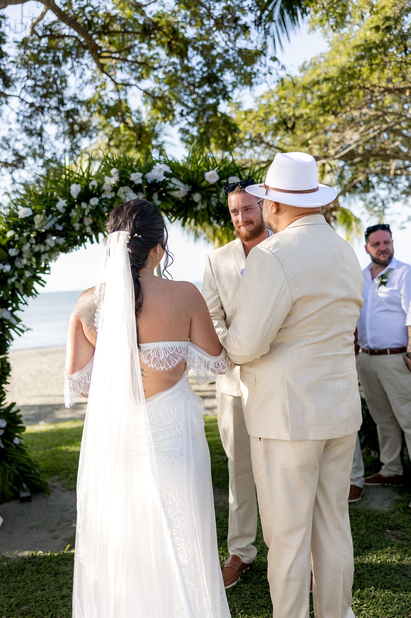 Bride and groom exchanging vows at outdoor beach wedding ceremony beneath floral arch with officiant.