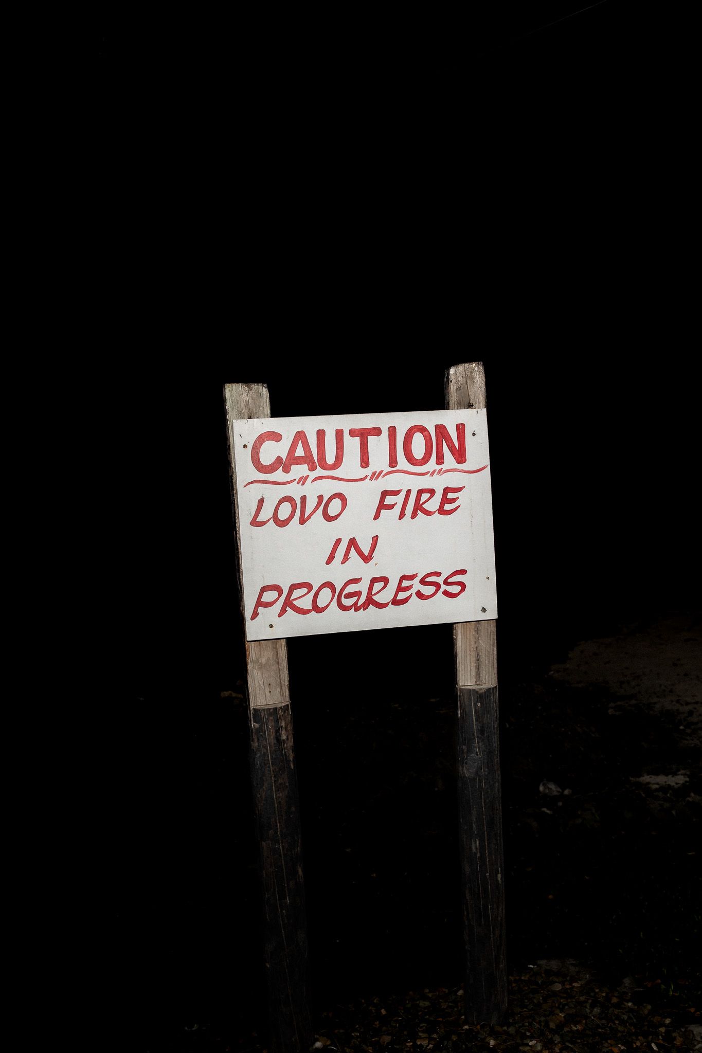 Caution sign warning of Lovo fire in progress mounted on wooden posts against dark background.