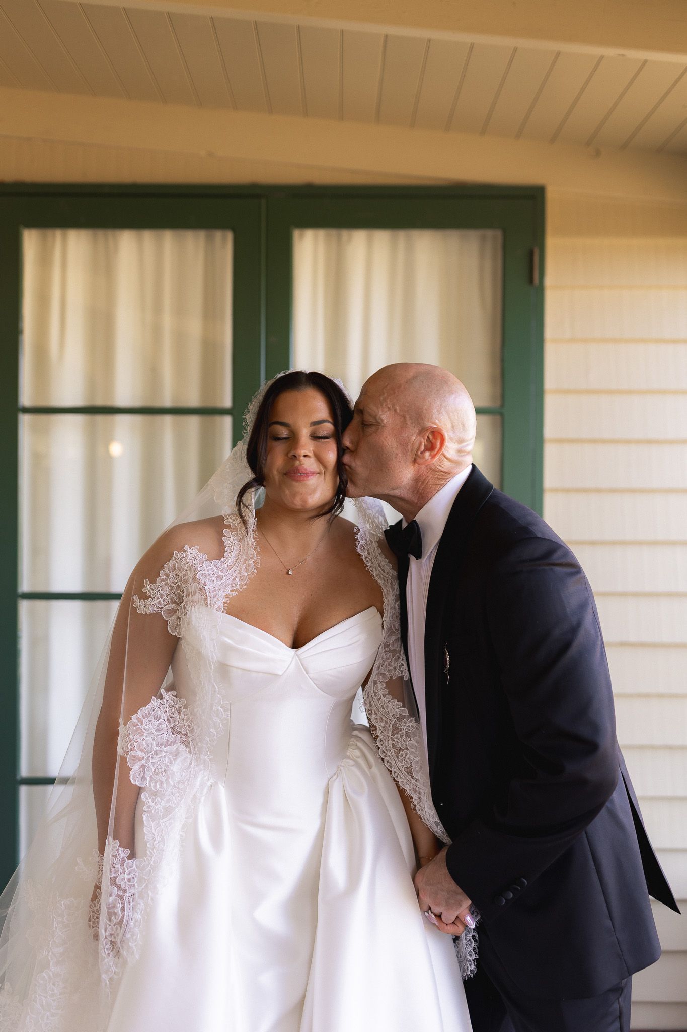 Bald groom in tuxedo kisses bride on cheek as she smiles in white lace wedding gown outdoors.
