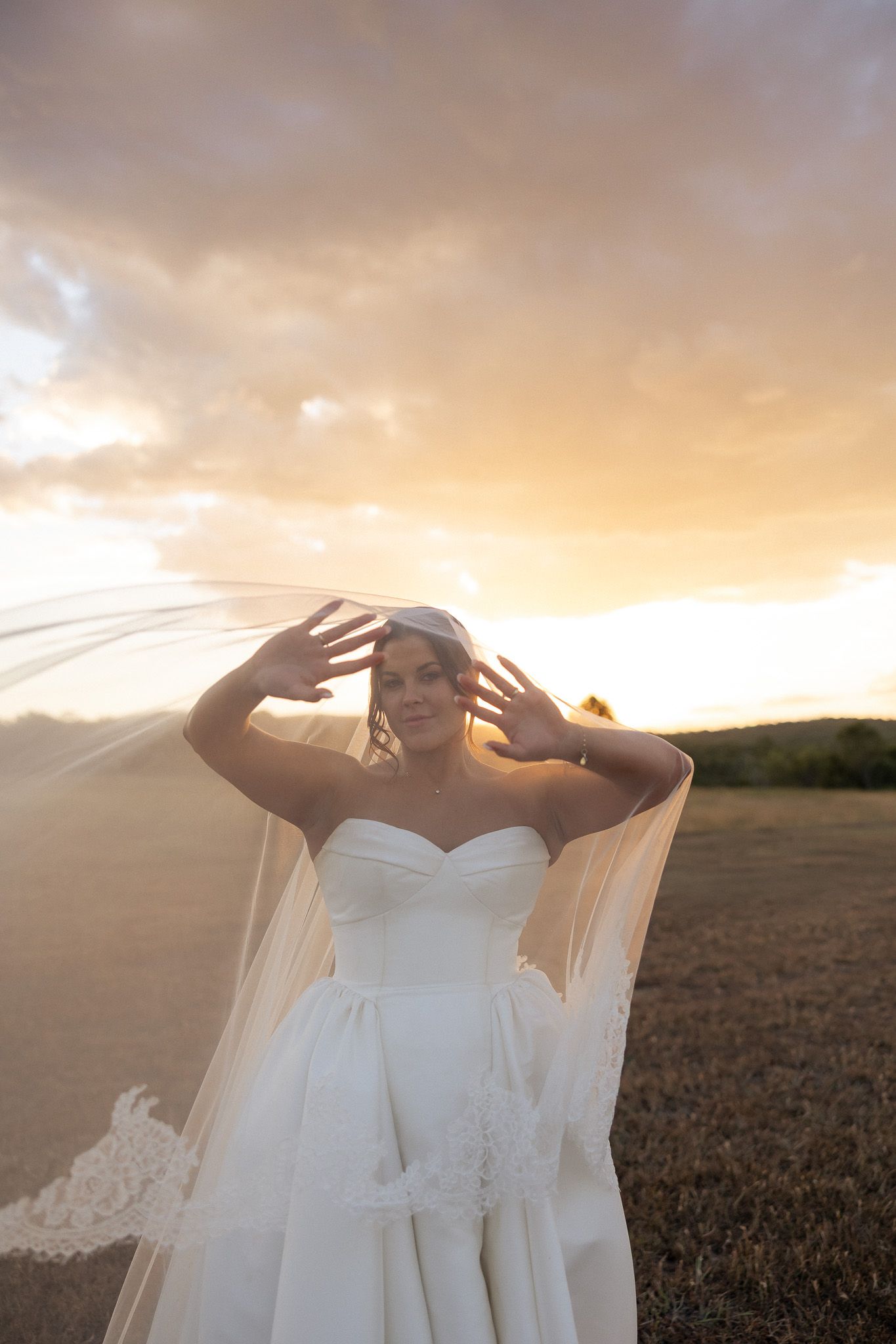 Bride in strapless white gown and lace veil poses at sunset in open field with dramatic golden sky.
