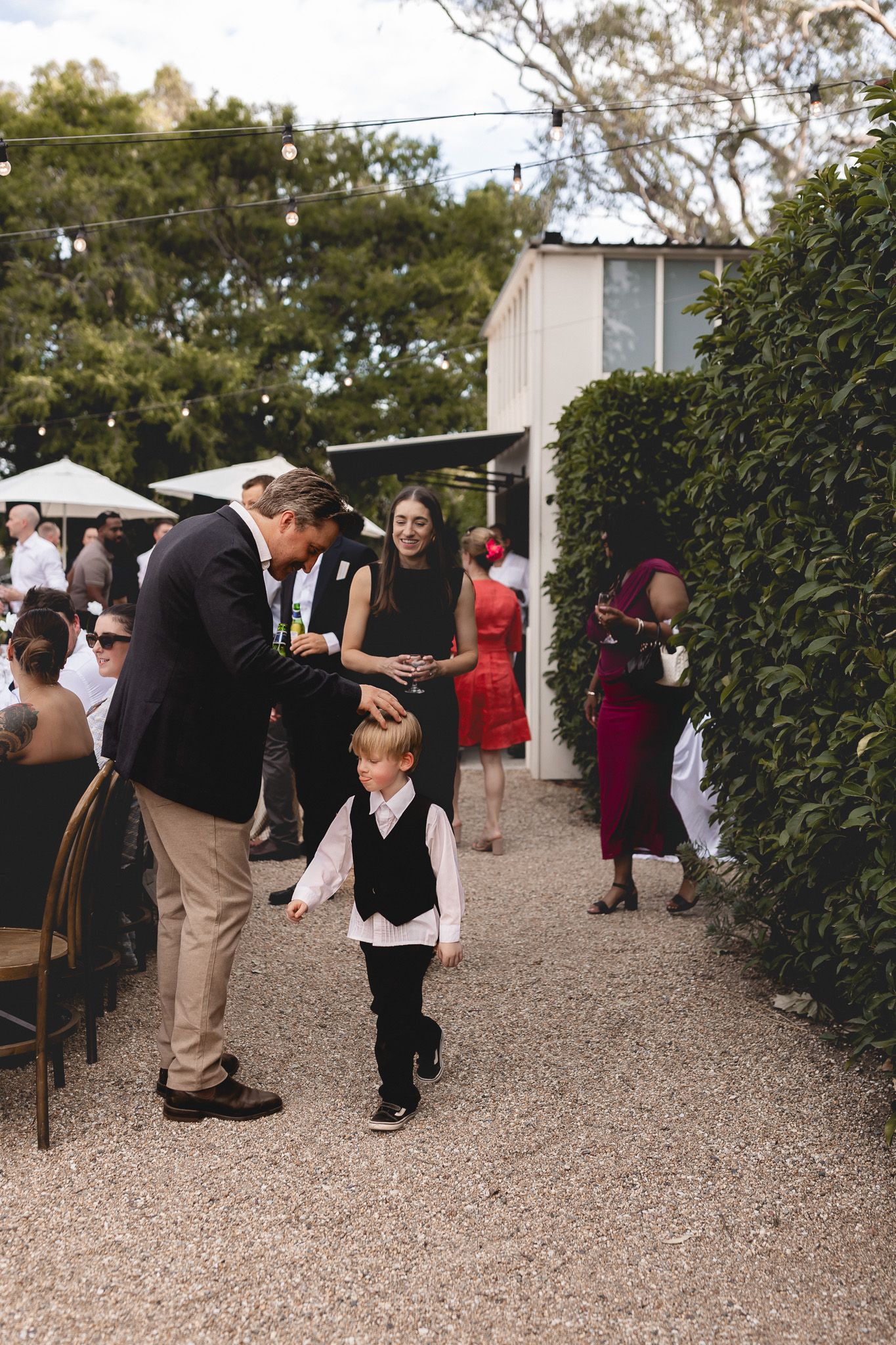 Wedding guests mingling outdoors with a young boy in a black vest walking along a garden pathway.