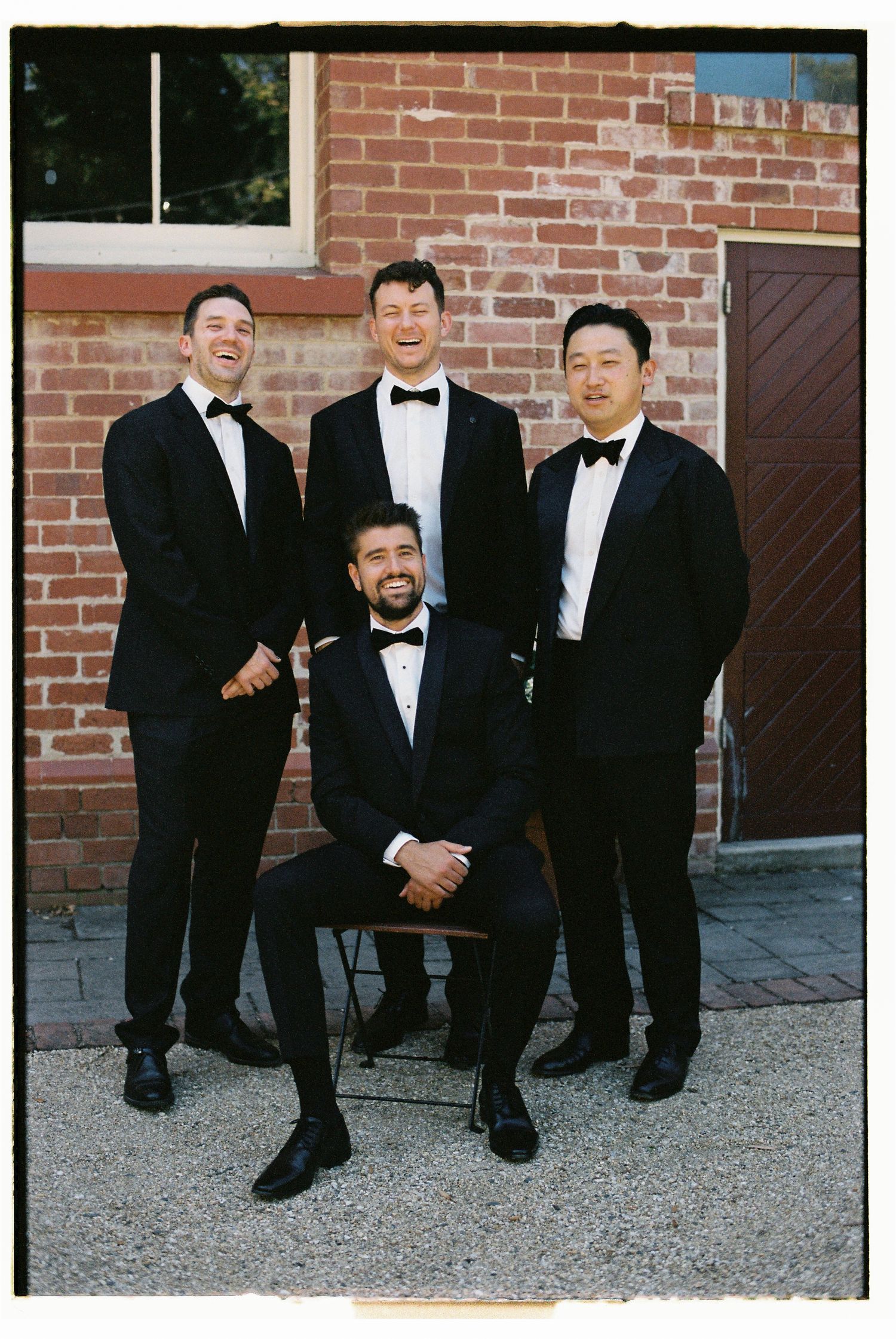 Four groomsmen in black tuxedos pose together outside a brick building, one seated and three standing behind him.
