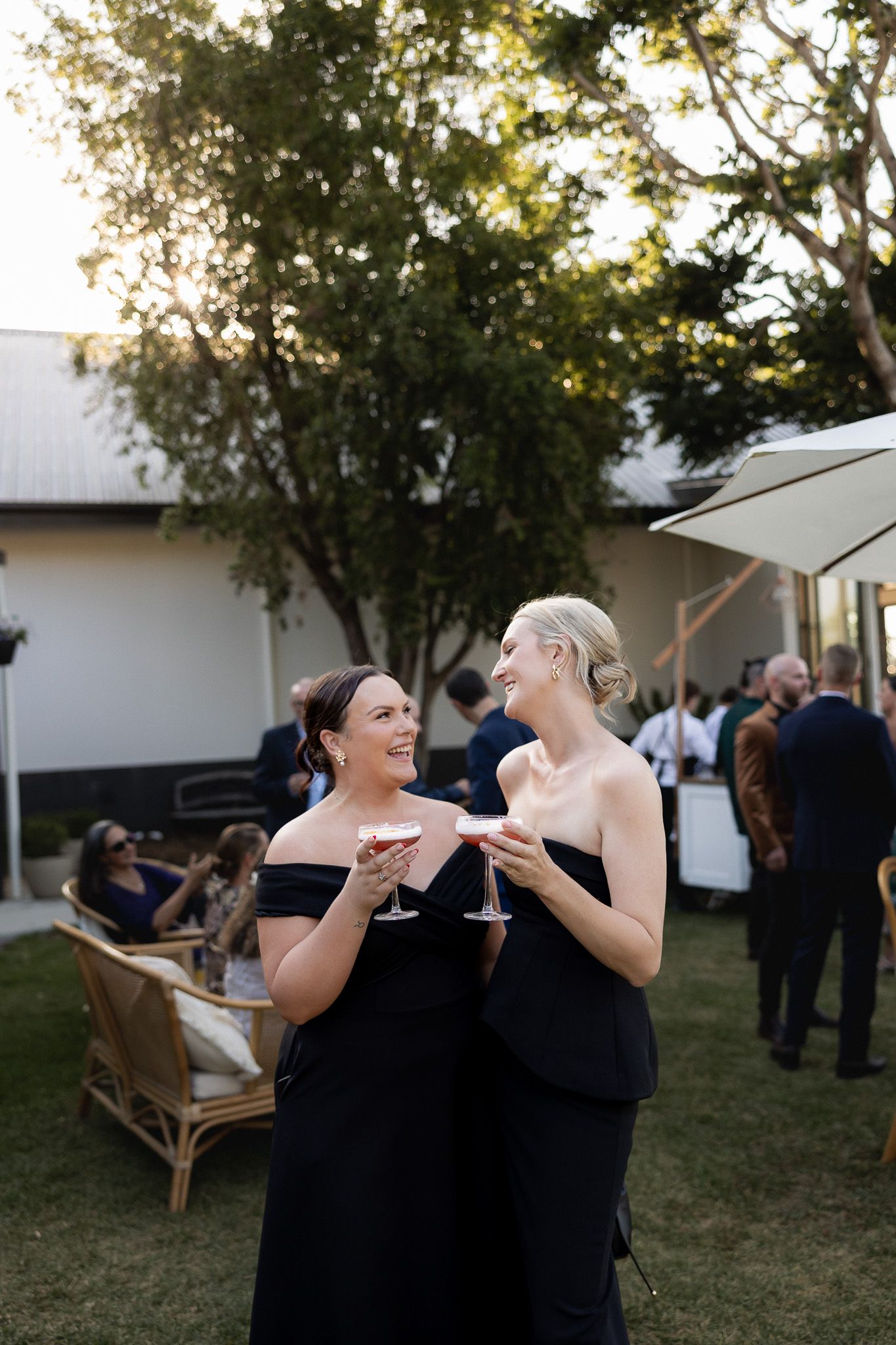 Two bridesmaids in black off-shoulder gowns laughing and holding cocktails at outdoor reception.