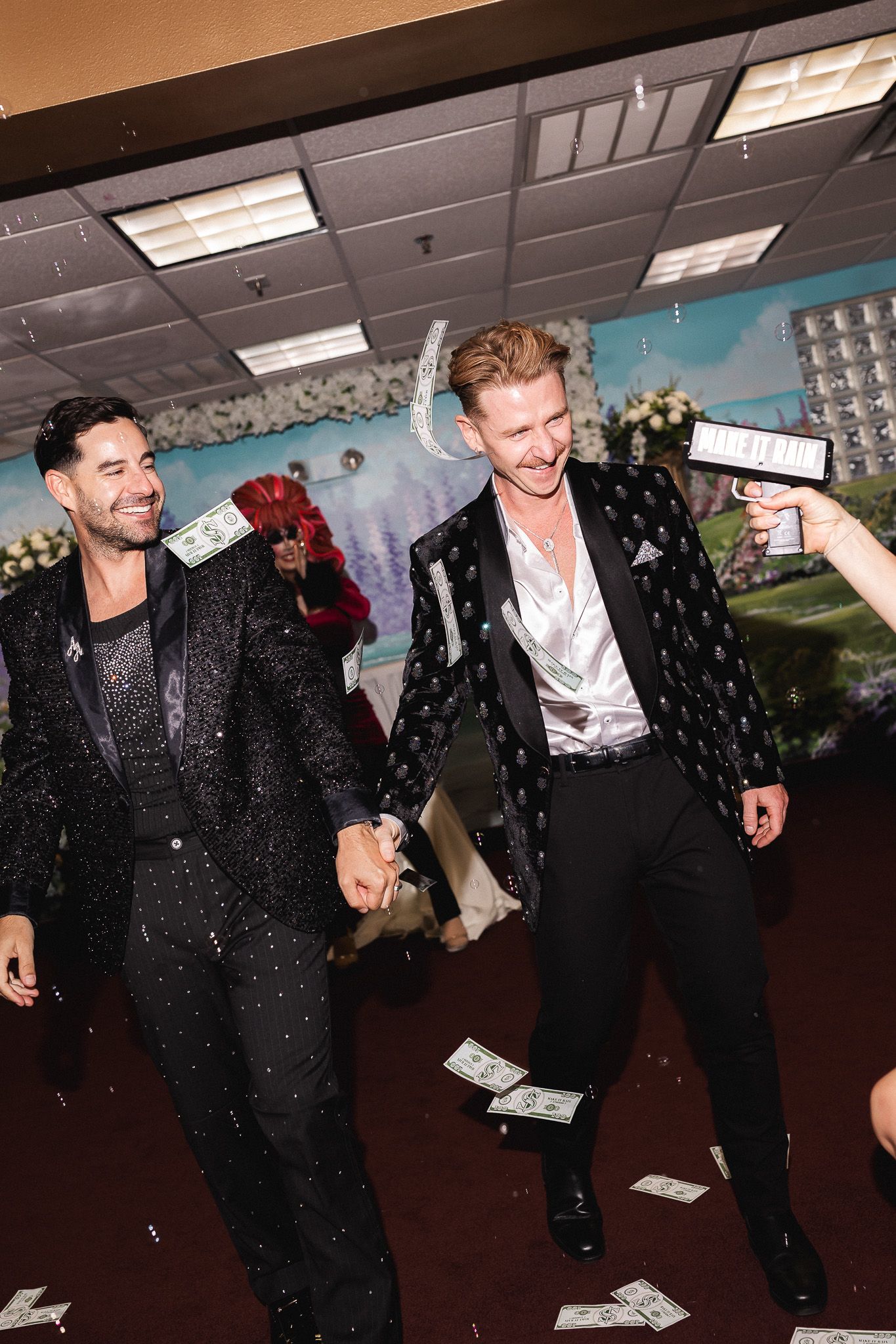 Two men in elegant black suits walk hand-in-hand as dollar bills rain down during a festive Las Vegas wedding ceremony.