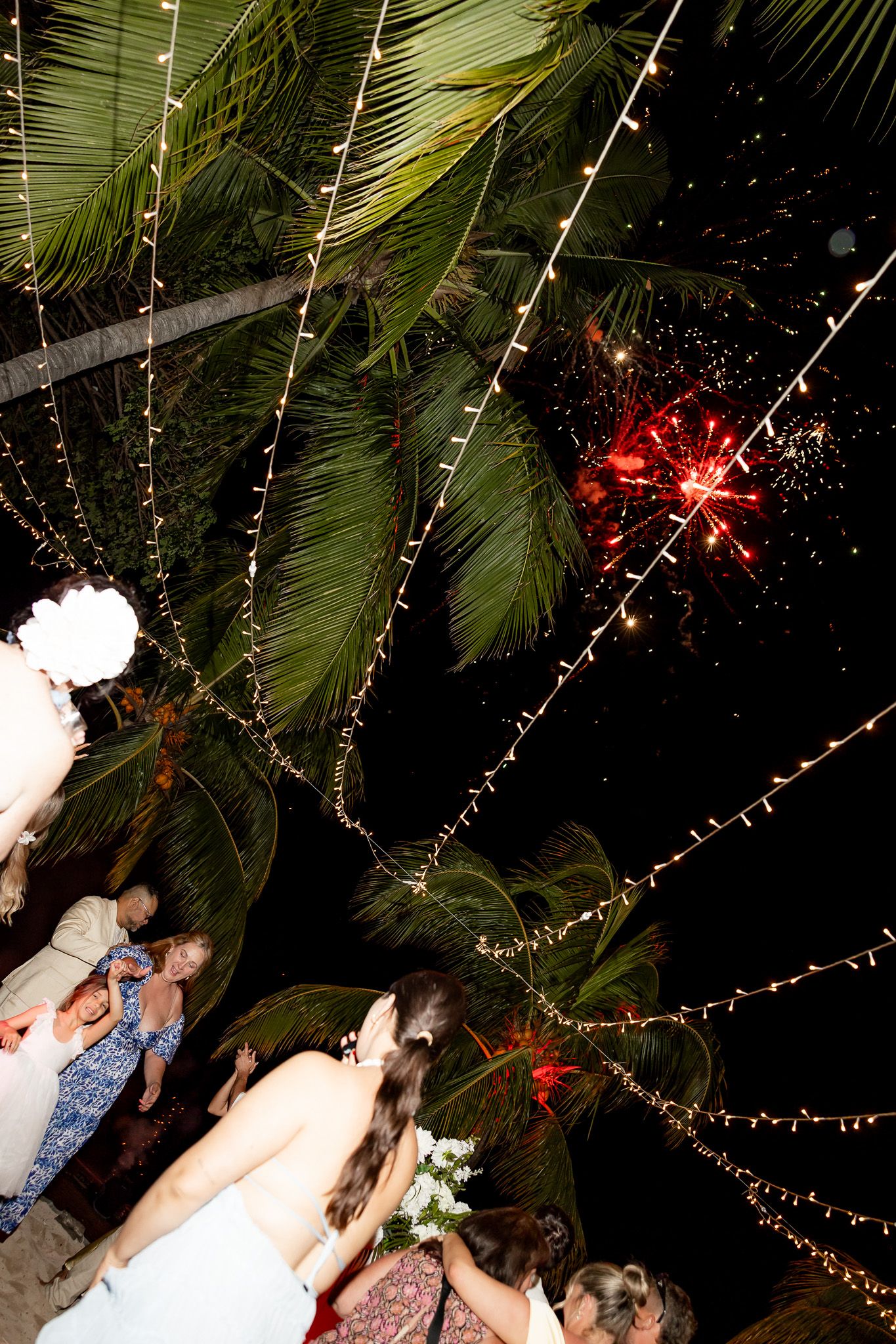 Wedding guests watching red fireworks burst above palm trees decorated with fairy lights at night reception.