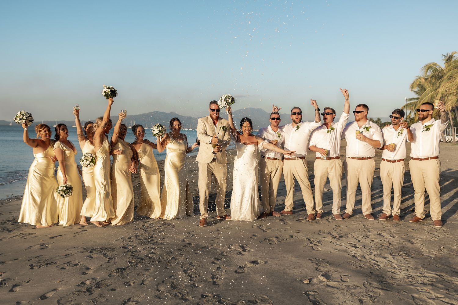 Wedding party celebrating on a beach at sunset, bridesmaids in ivory gowns and groomsmen in white shirts raising hands joyfully.