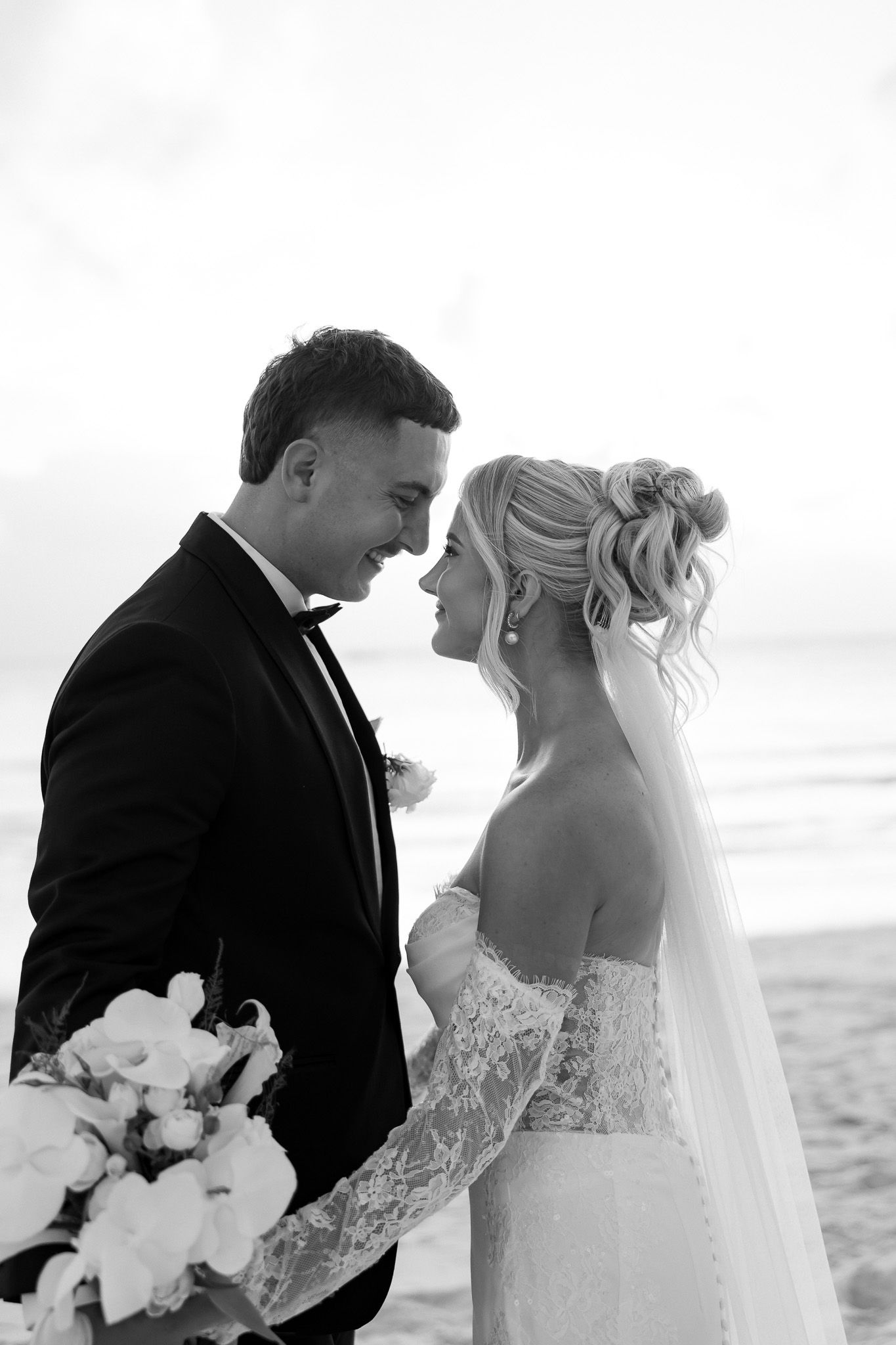 Black and white photo of a bride and groom sharing an intimate moment on a beach, bride holding a bouquet.