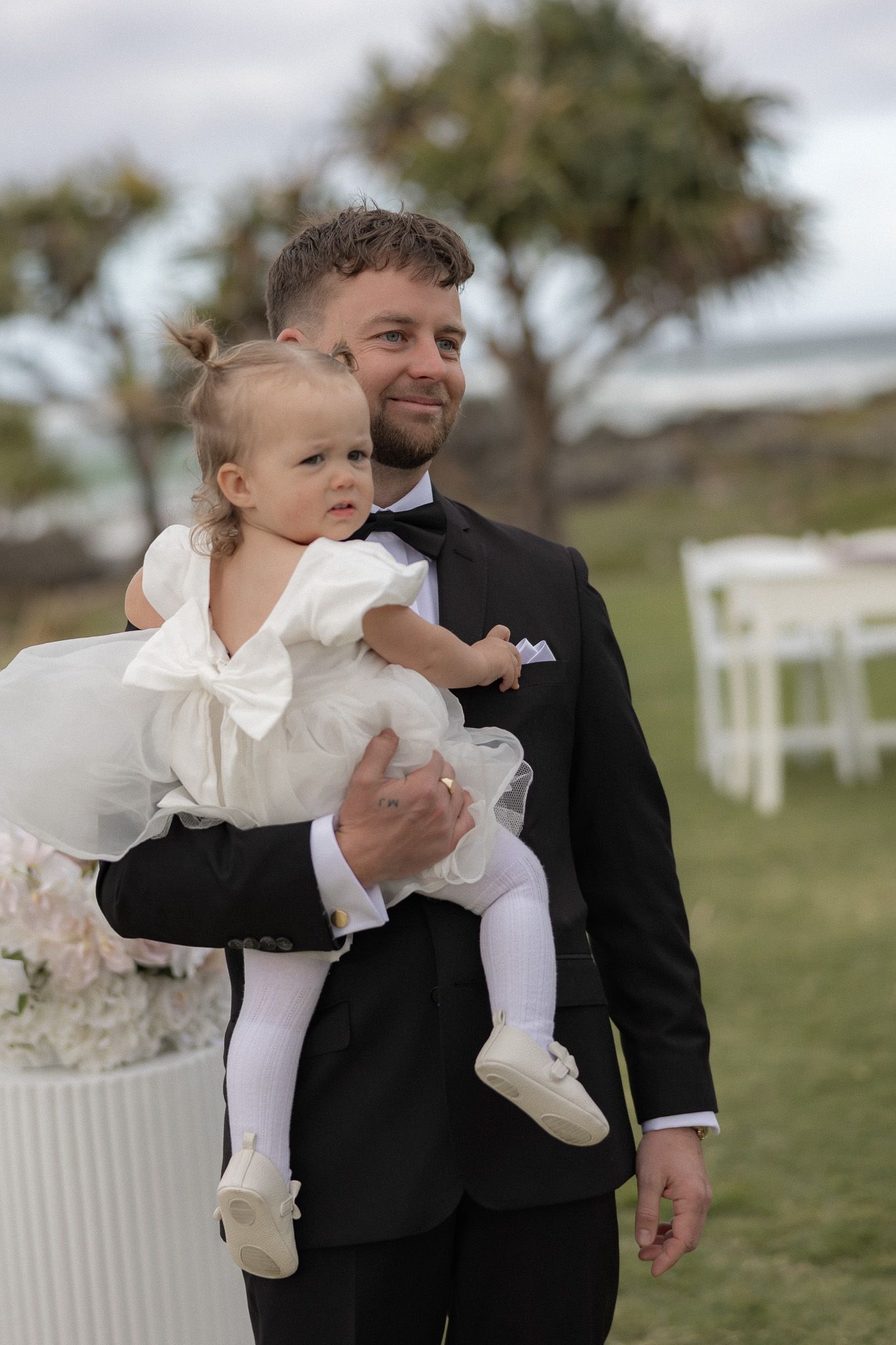 Groom in black tuxedo holding baby girl in white dress at outdoor wedding ceremony.