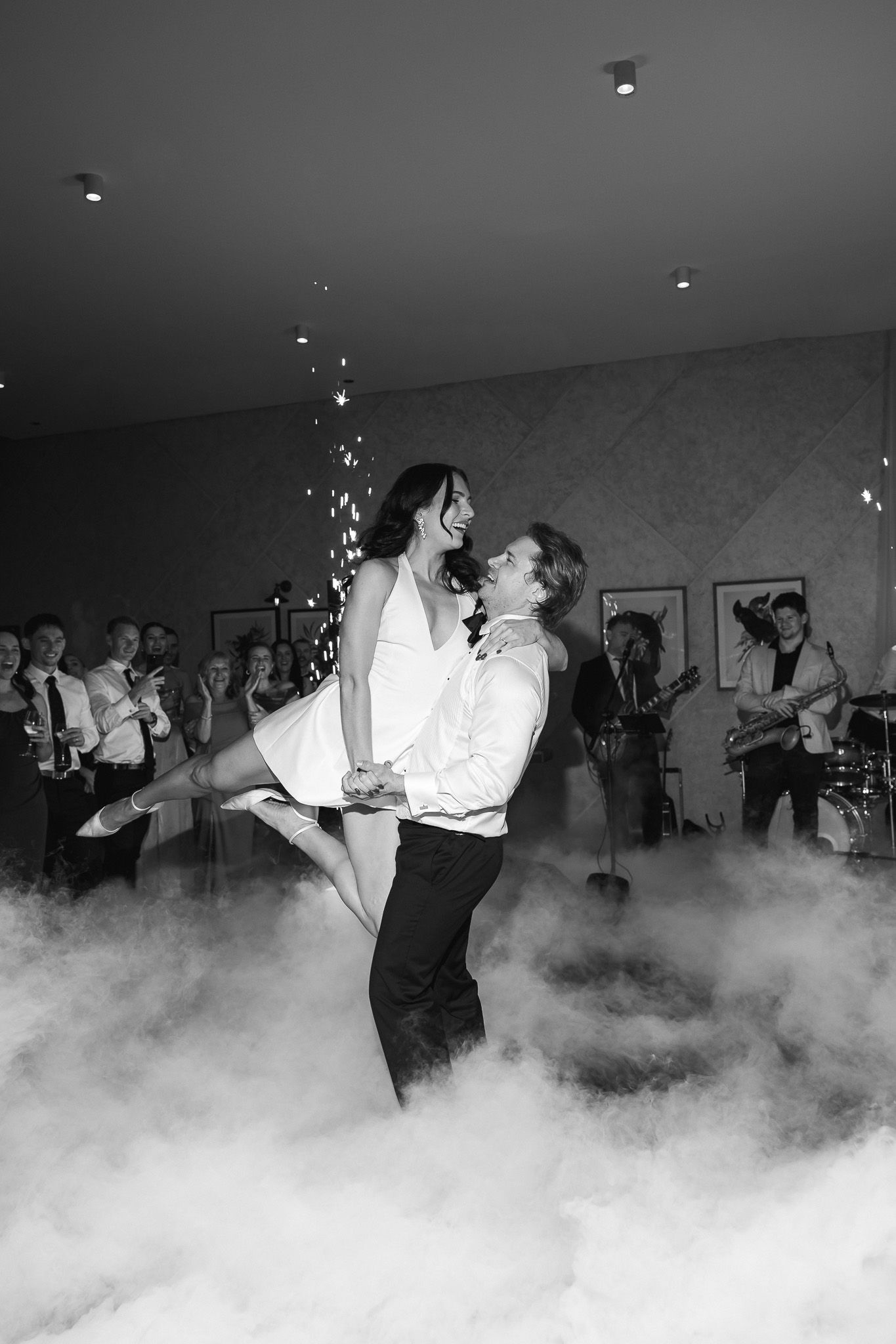 Groom lifting bride during first dance surrounded by fog and wedding guests in black and white photo.