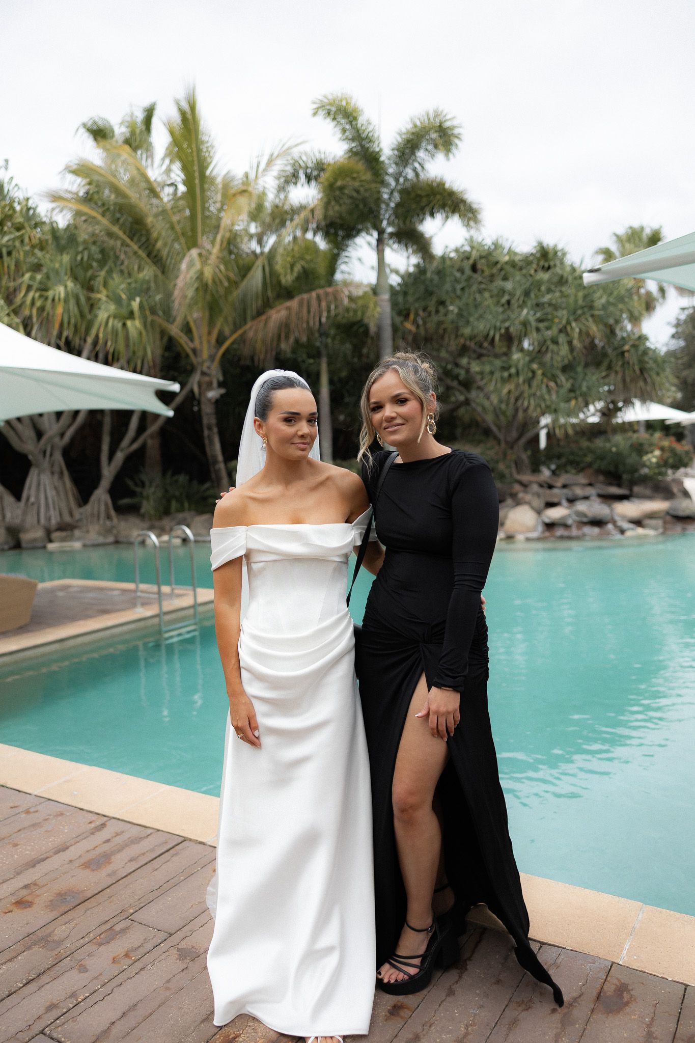 Bride in off-shoulder white gown and veil poses with guest in black dress by pool.