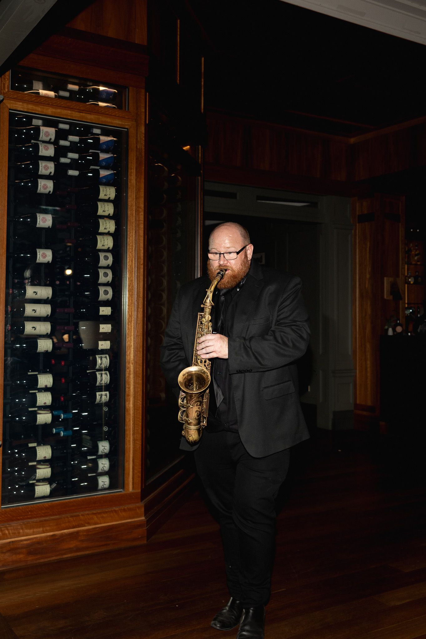 Bearded saxophonist in black suit playing alto saxophone beside a wine cellar wall at an elegant event.