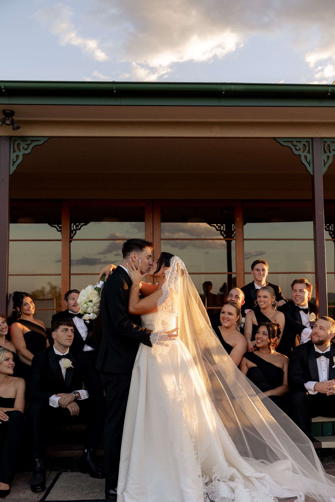 Bride and groom share a kiss in golden sunset light with wedding party gathered on venue porch.