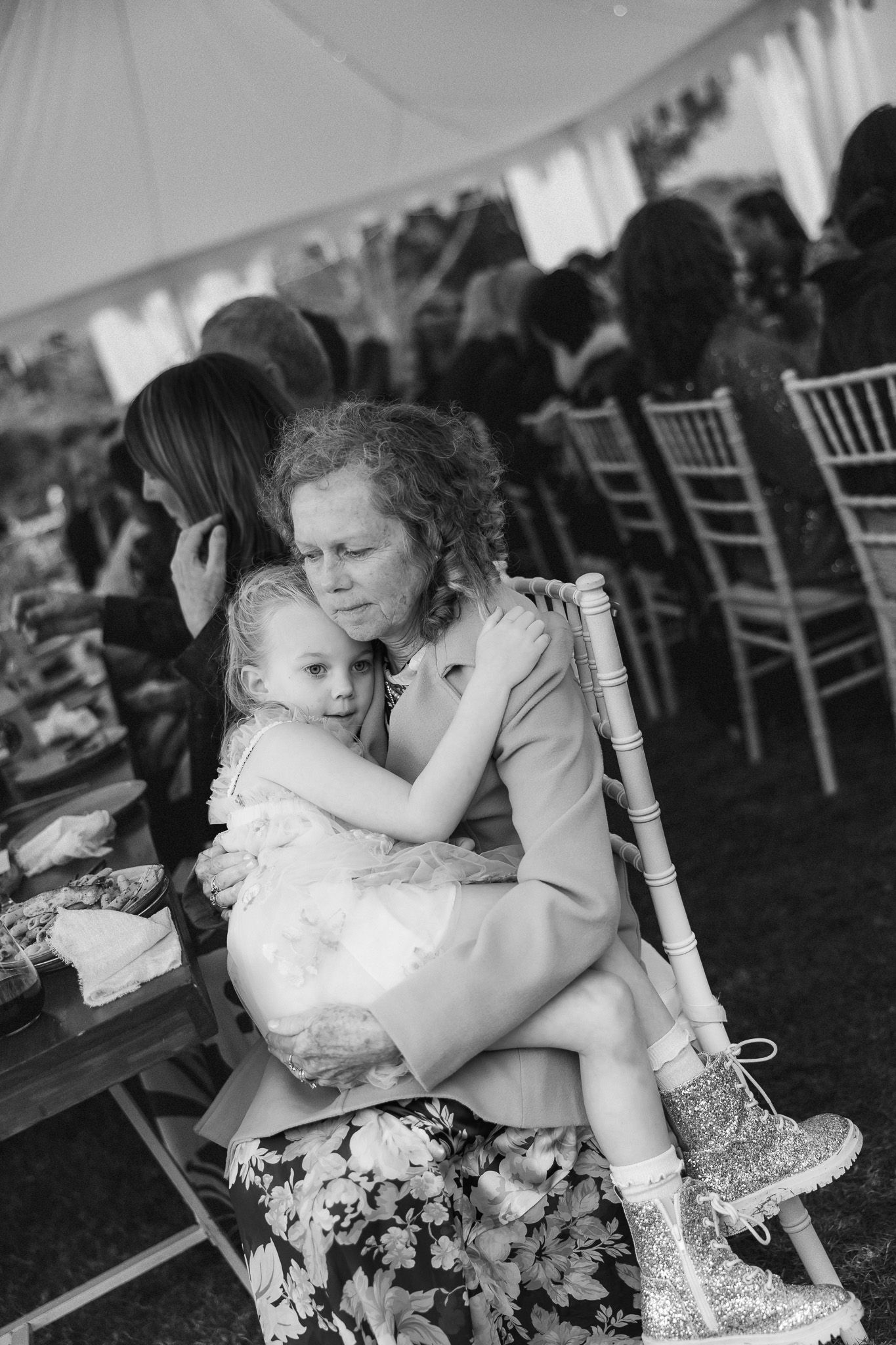 Black and white photo of a grandmother hugging a young girl in sparkly shoes at a wedding.
