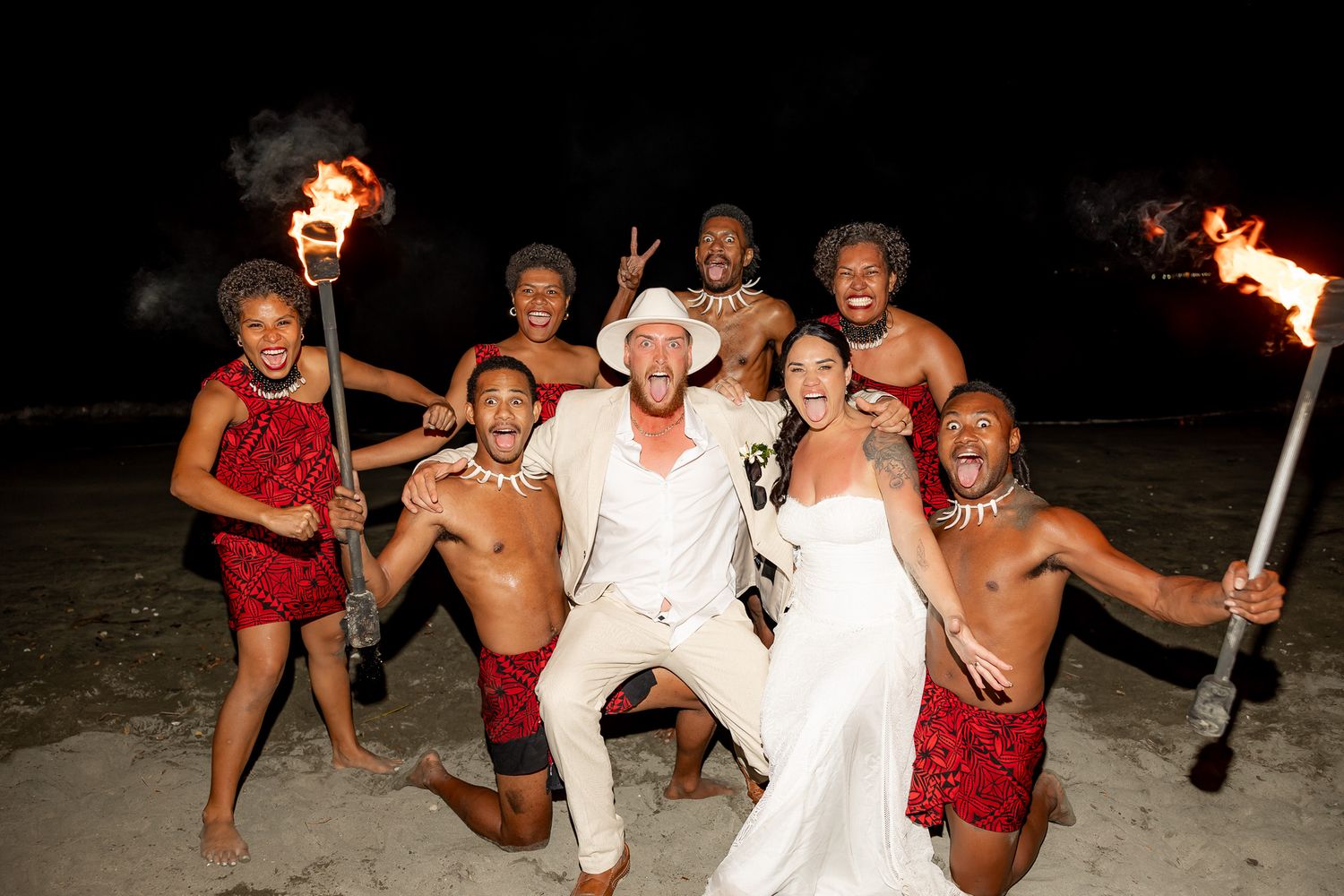 Bride and groom posing with fire dancers in red costumes on beach at night during wedding celebration.
