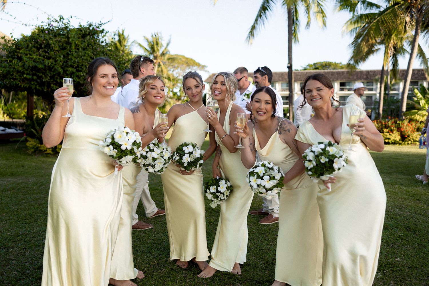 Bridesmaids in yellow satin dresses holding white bouquets and champagne glasses at a tropical outdoor wedding.