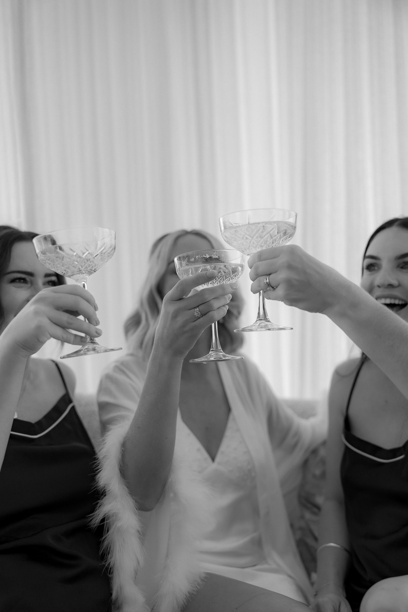 Black and white photo of bride and bridesmaids toasting with crystal champagne glasses while getting ready.