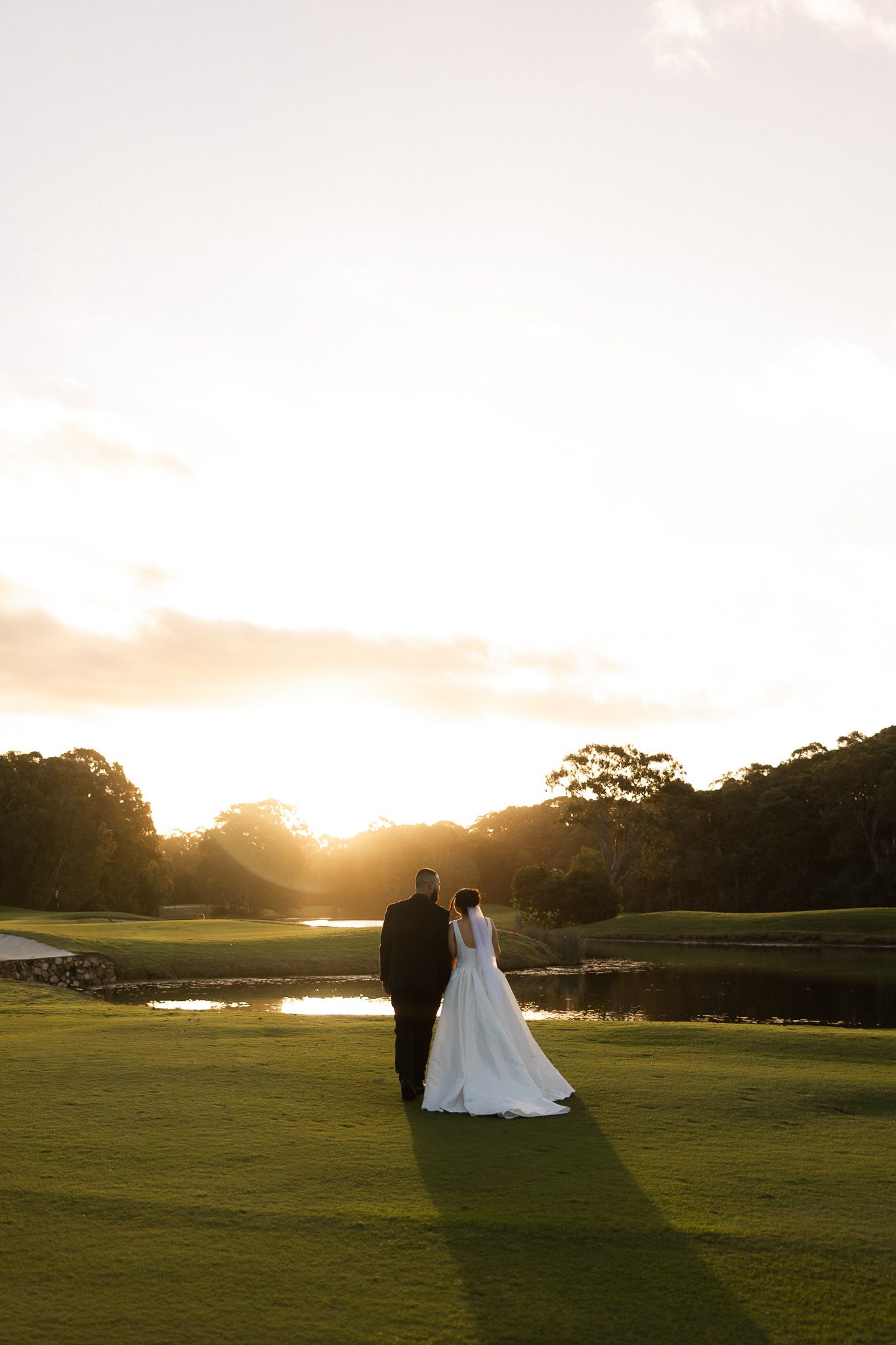 Bride and groom standing on lush green golf course at golden sunset, with pond and trees in background.