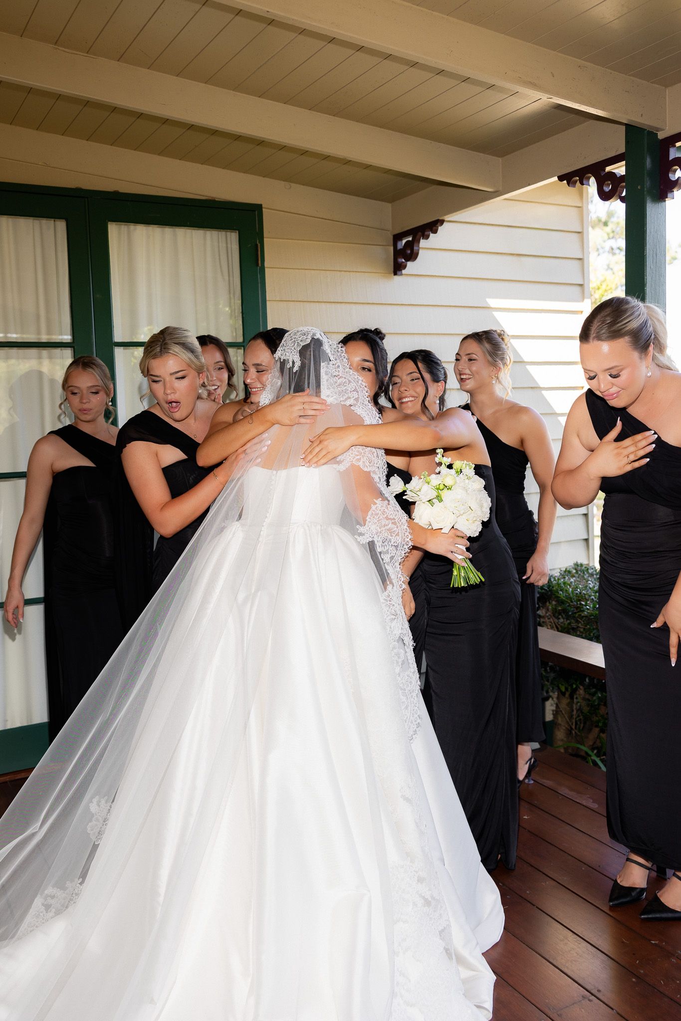 Excited bridesmaids in black dresses react with joy upon seeing bride in white ball gown and veil.