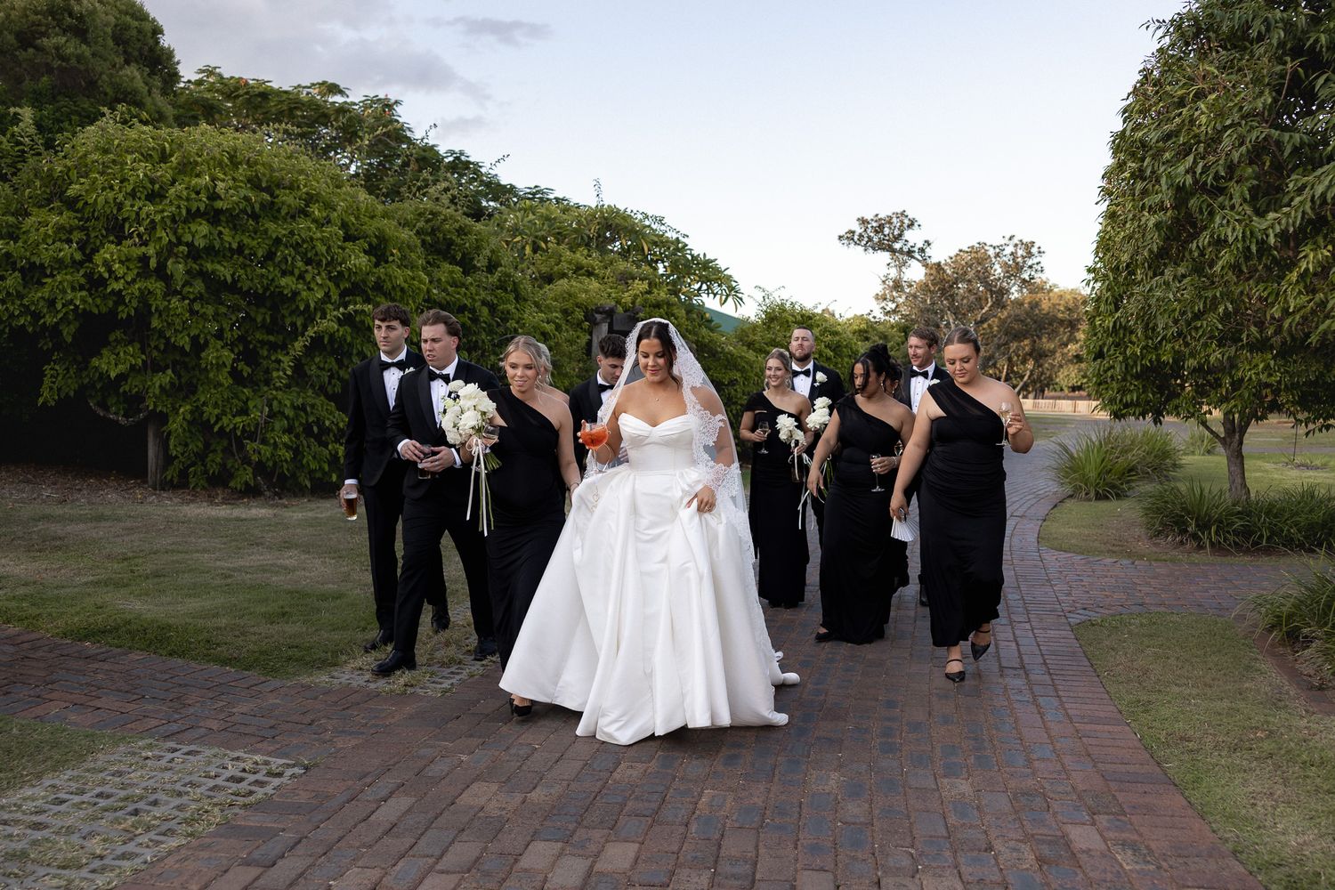 Bride in white ballgown leading wedding party in black attire along a brick pathway through lush garden grounds.