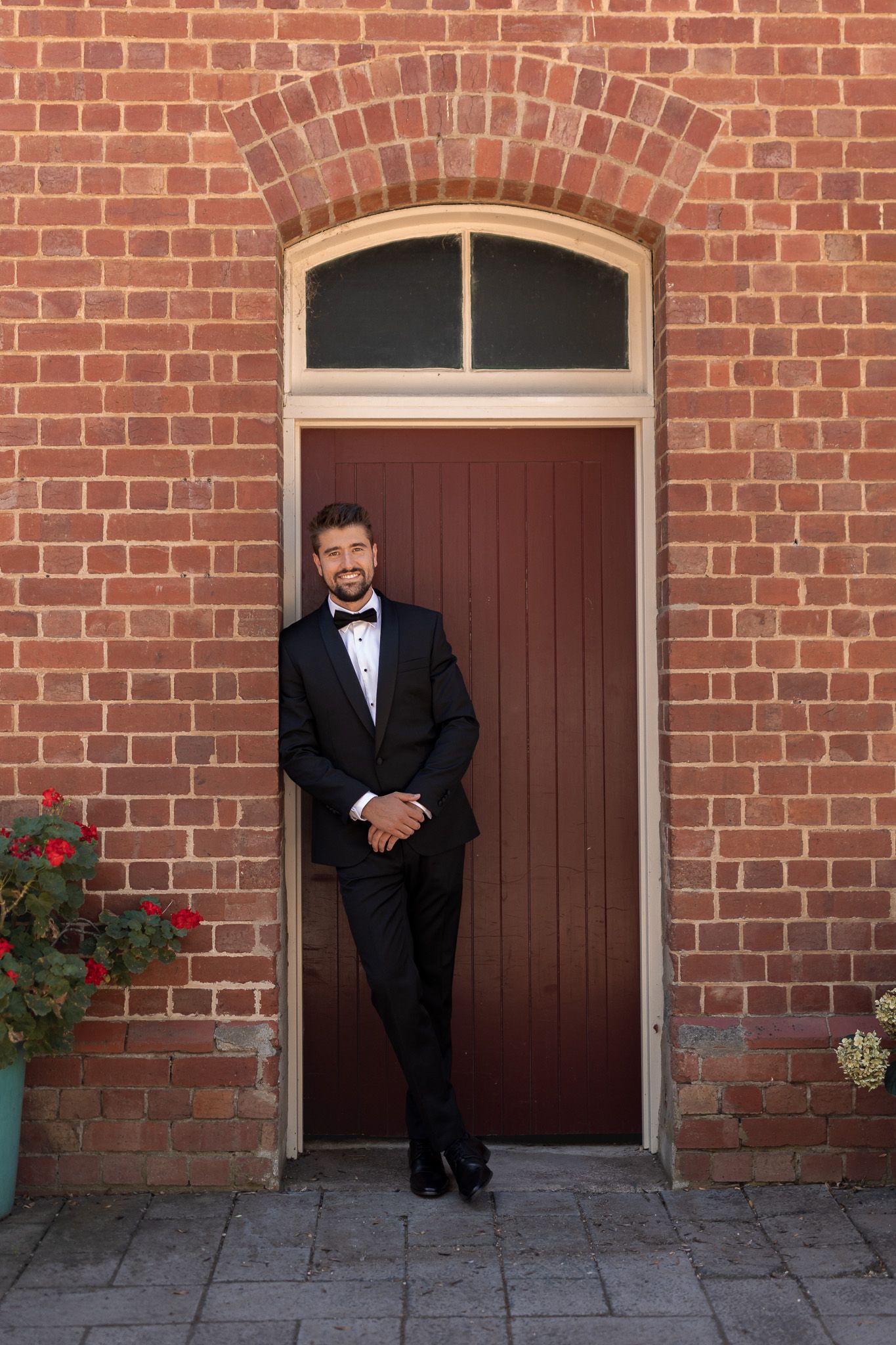 Handsome groom in black tuxedo with bow tie leaning against a red brick wall by a wooden door.