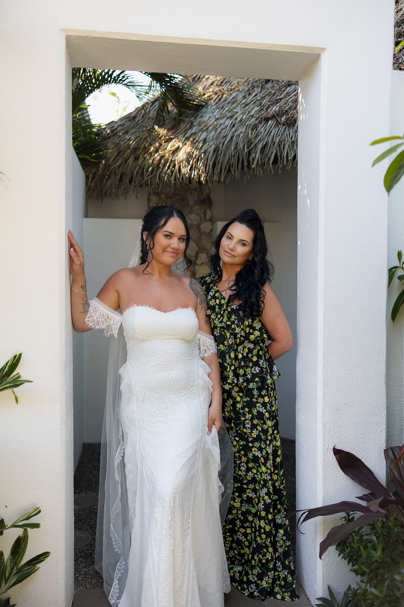 Bride in white lace gown posing with dark-haired woman in floral dress in white archway.