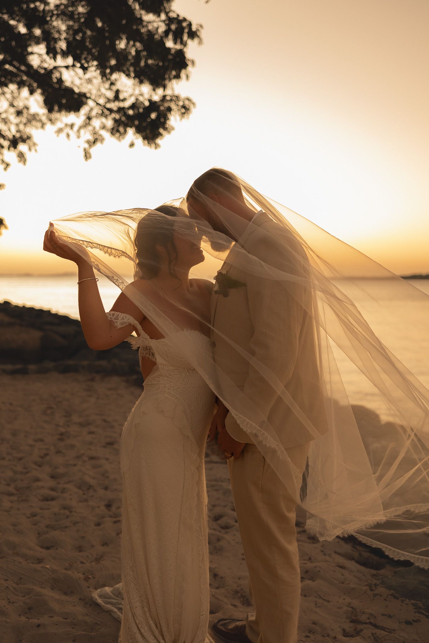 Bride and groom share a romantic moment under a flowing veil at a stunning beach sunset wedding.