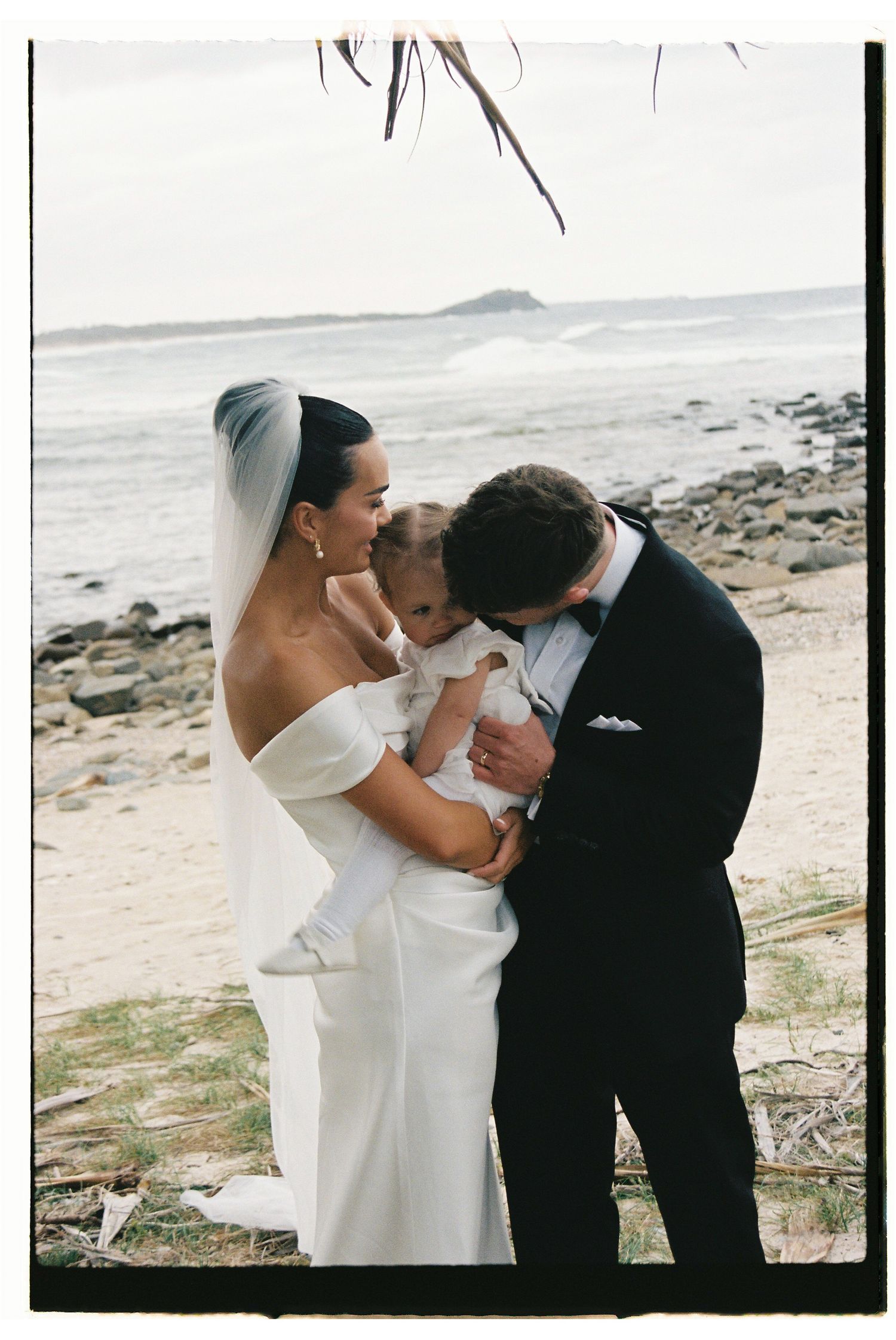 Bride in off-shoulder gown and groom in tuxedo share tender moment with baby on rocky beach.