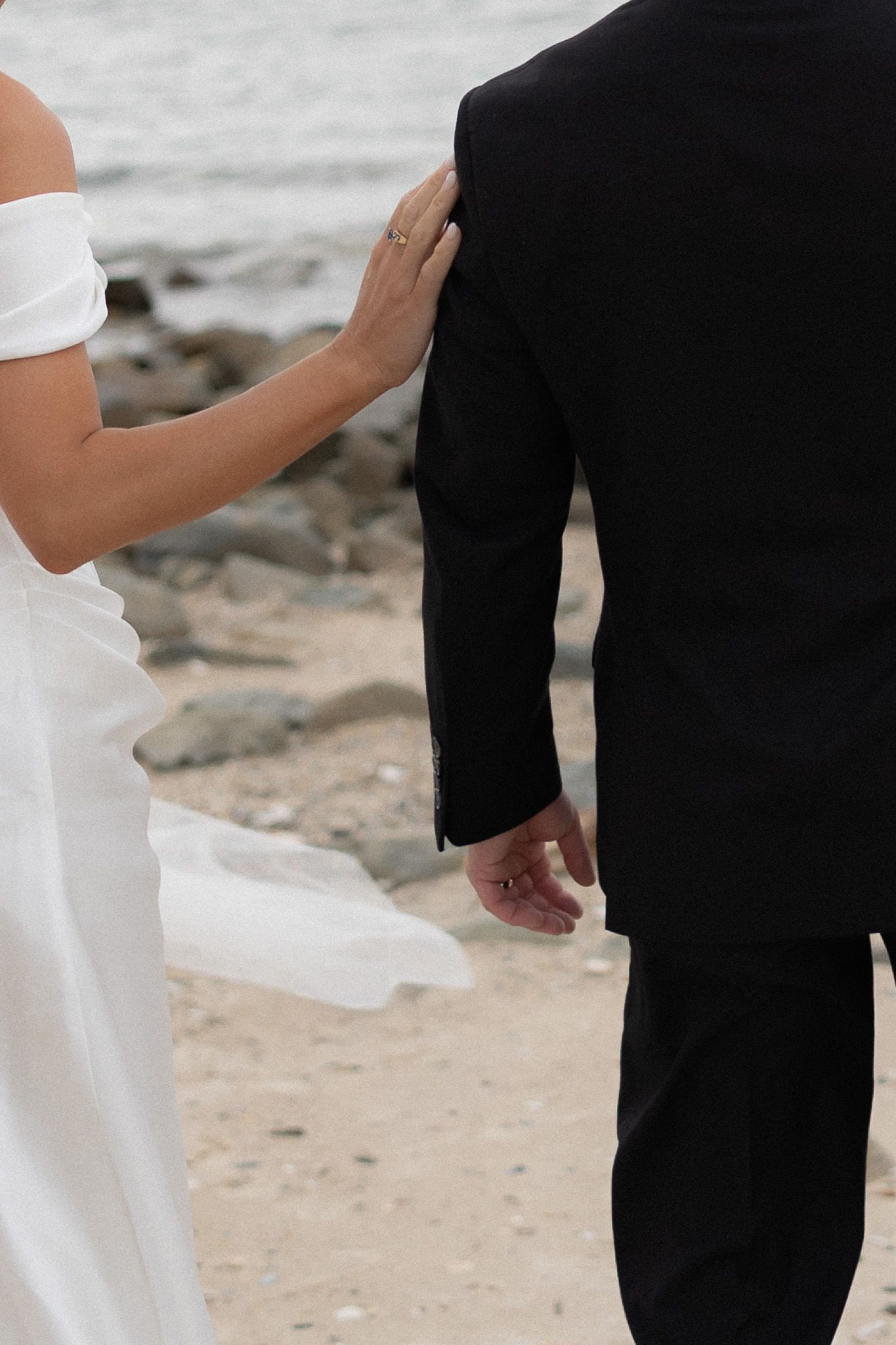 Bride in off-shoulder white gown places hand on grooms black suit jacket at beach.