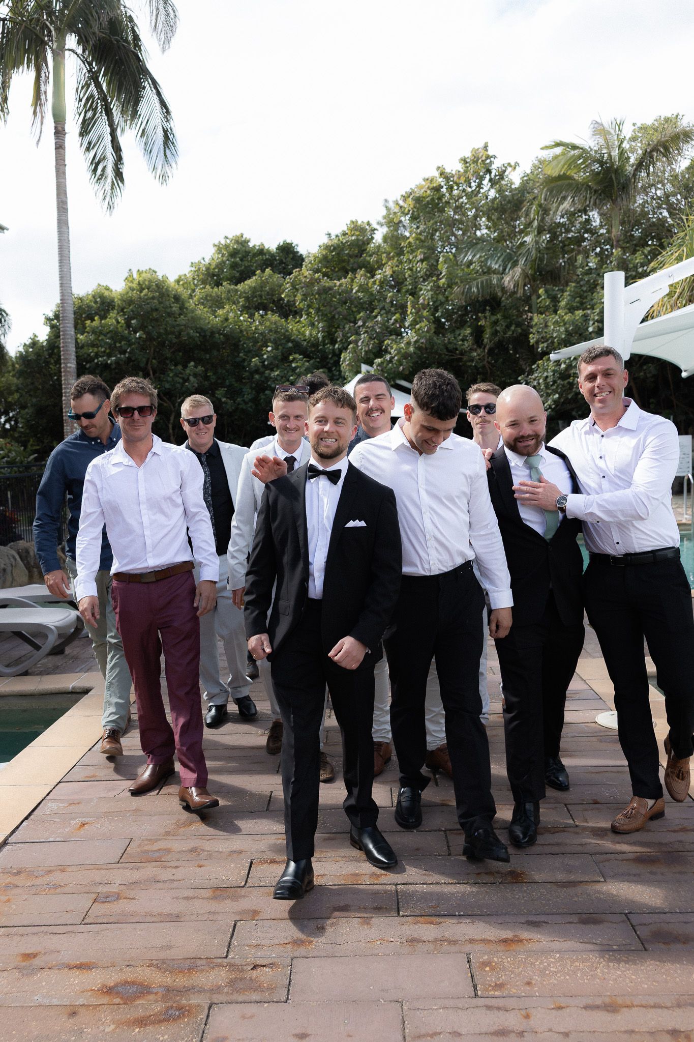 Groom in black tuxedo leads groomsmen in white shirts walking poolside at outdoor wedding venue.