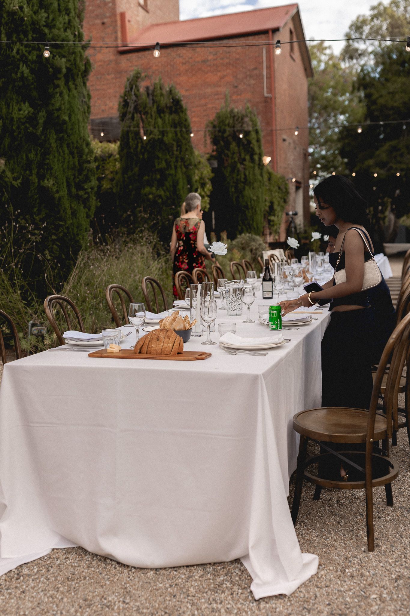 Elegant outdoor dining table set with white linen, glassware, and bread board, with guests preparing for an al fresco event.