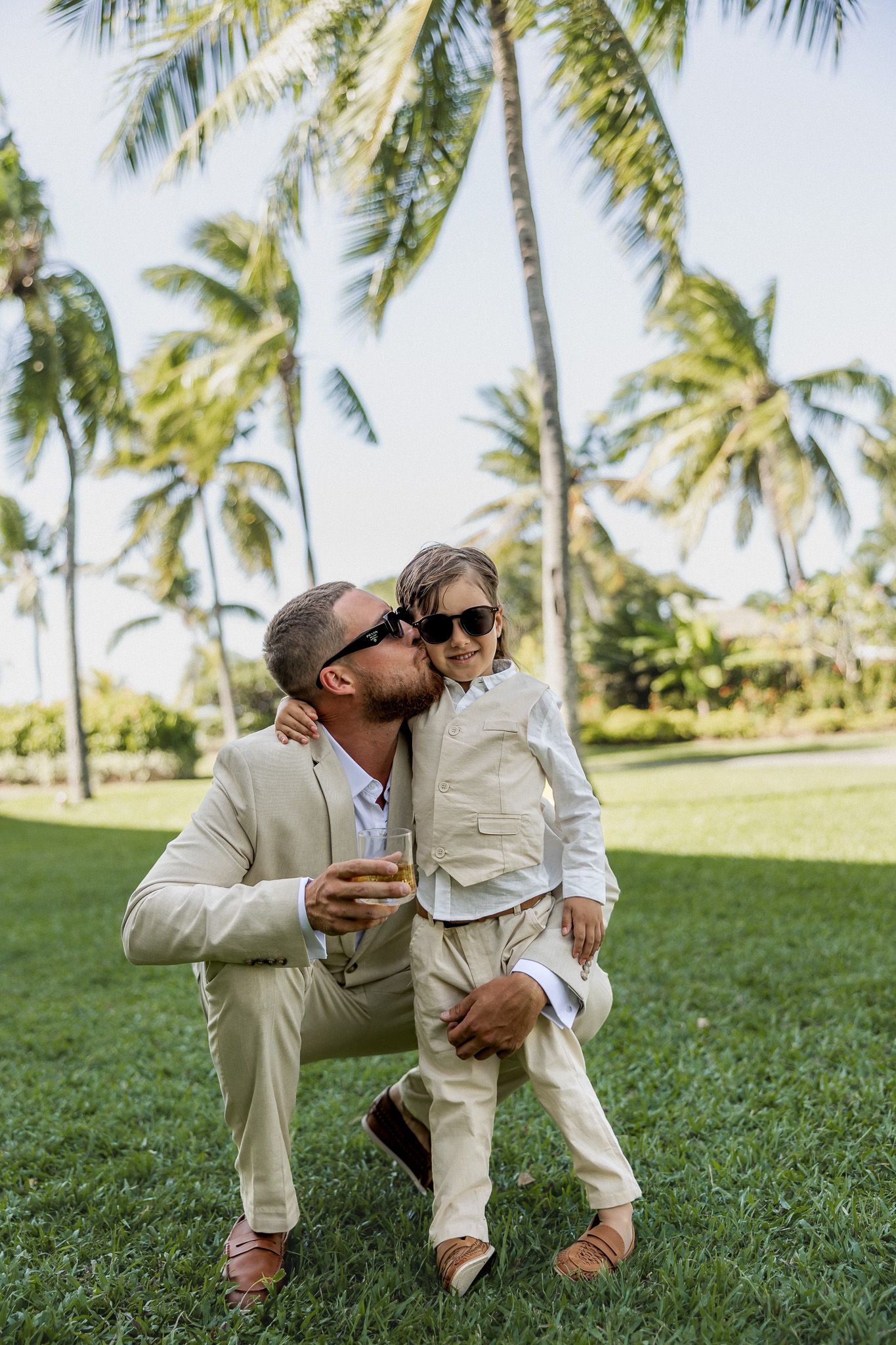Father in beige suit kissing young child in sunglasses on grass with palm trees behind.