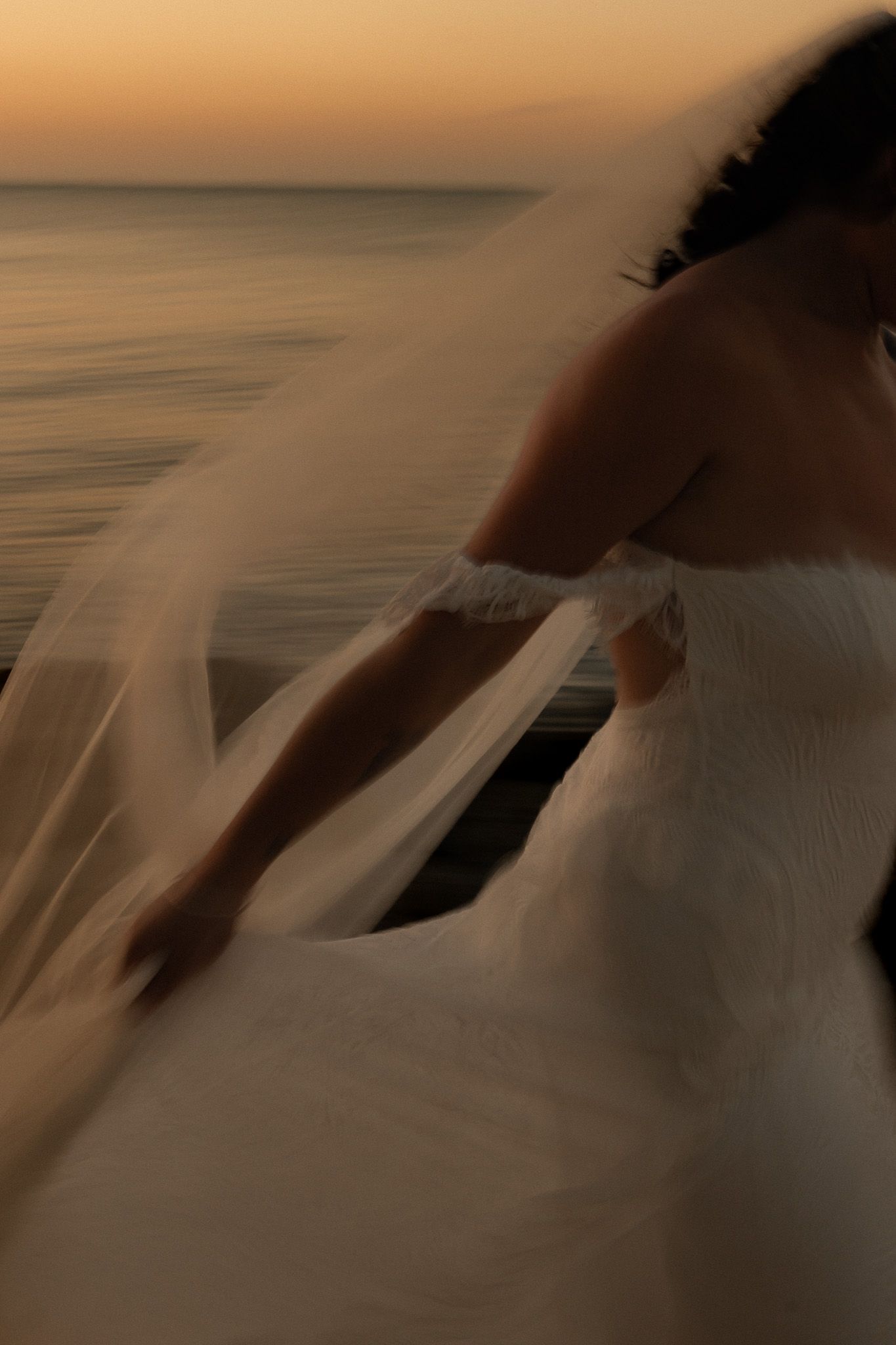 Bride in white lace gown with flowing veil stands near water at golden sunset.
