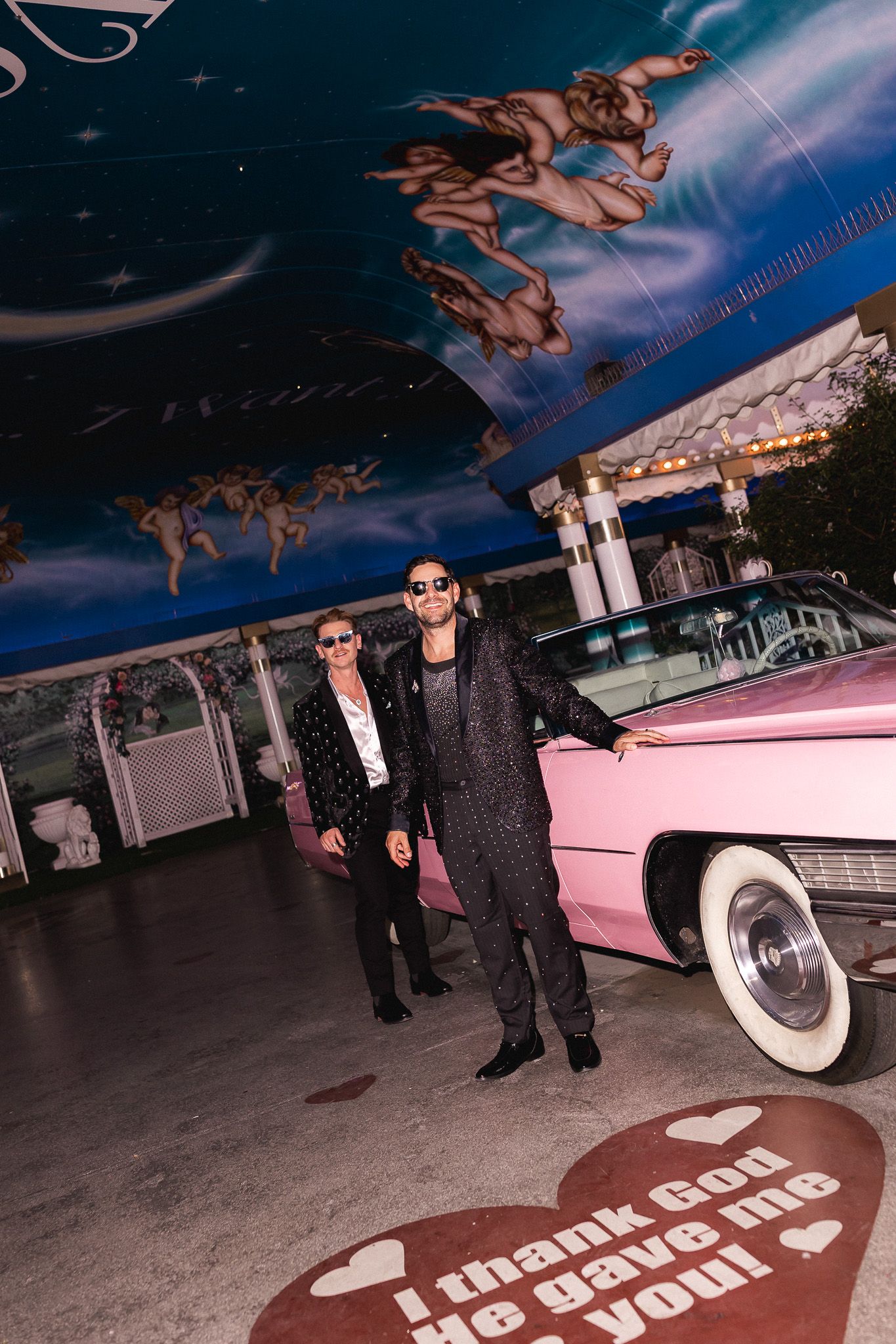 Two people in sparkly outfits pose beside a classic pink Cadillac under a celestial painted ceiling.