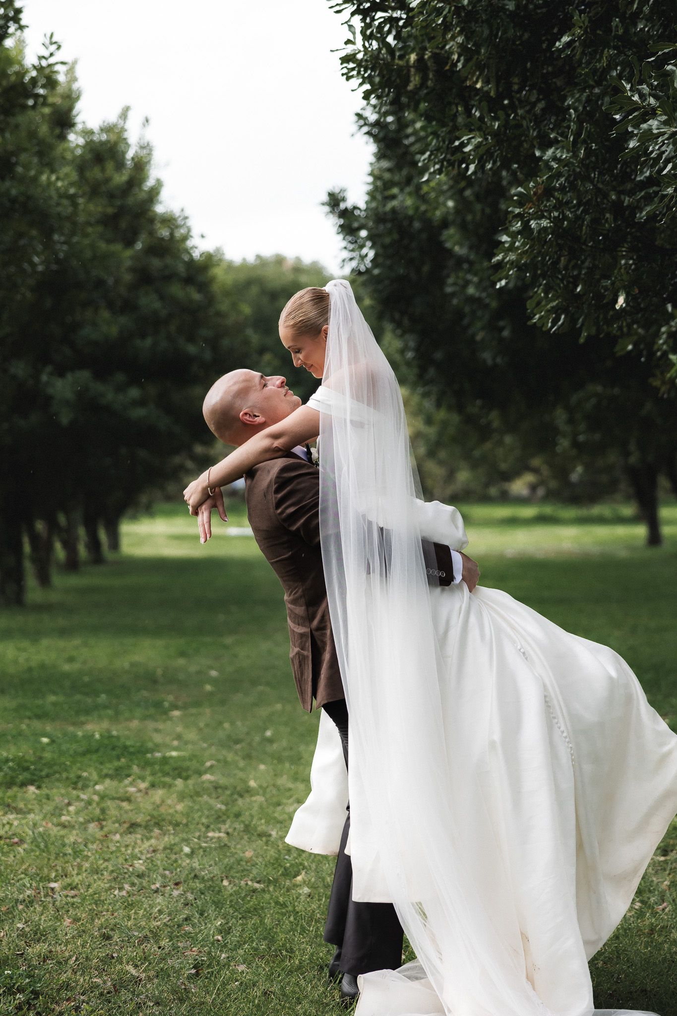 Groom lifting bride in white wedding gown and veil in a lush green park setting.