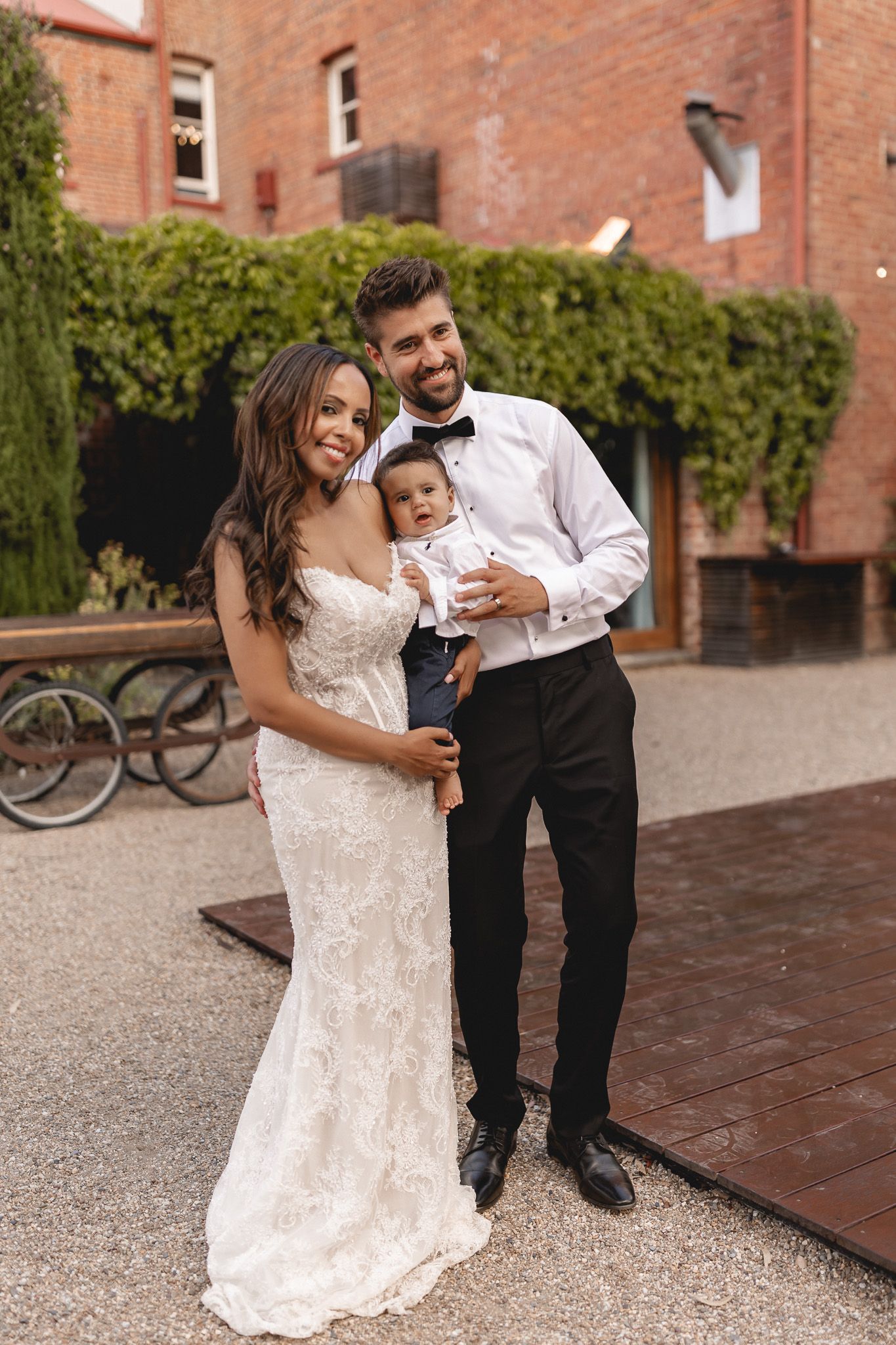 Smiling bride and groom in lace gown and black tuxedo hold their baby at outdoor venue.