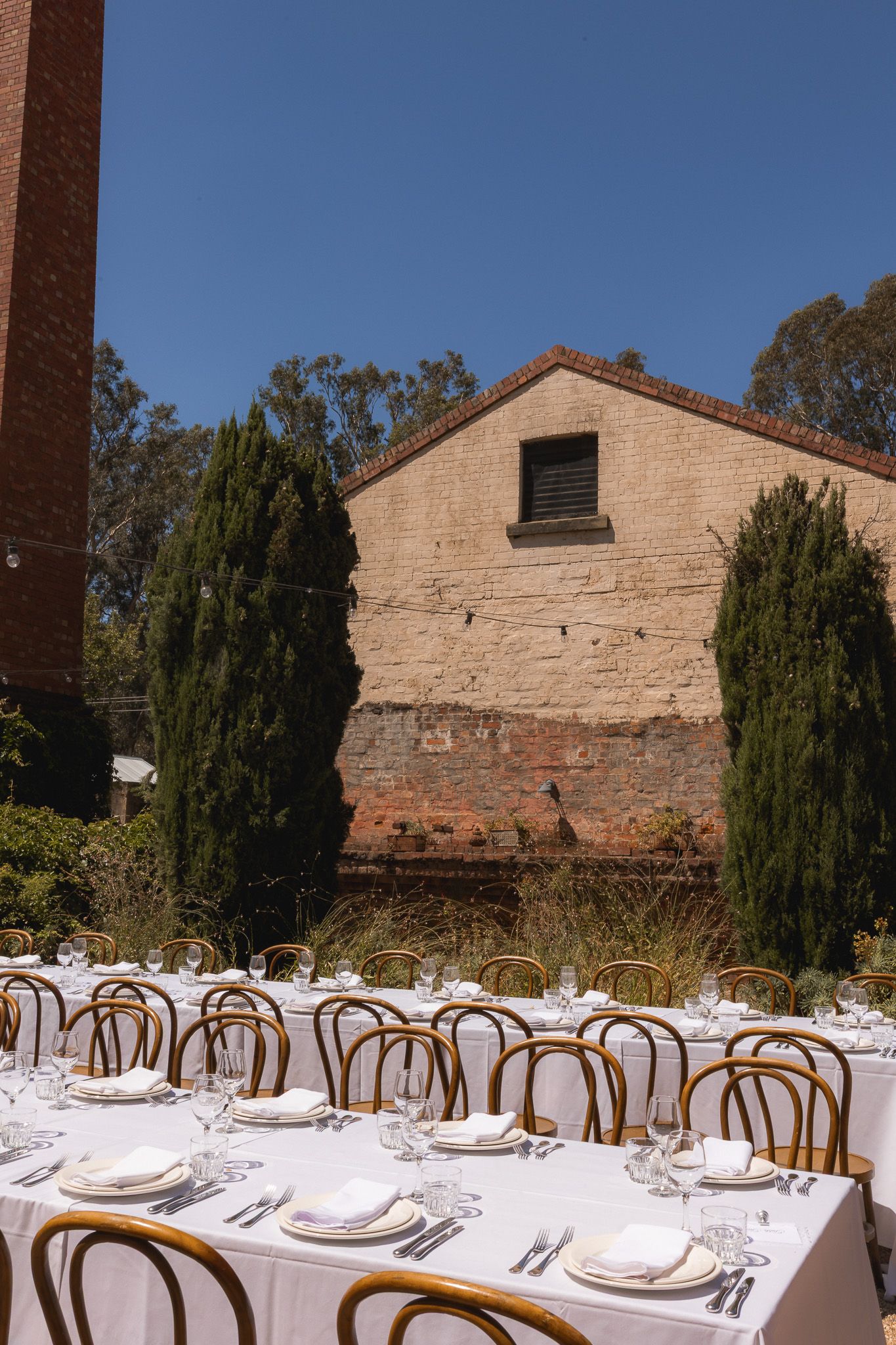 Outdoor wedding dinner table set with white plates and glassware, bentwood chairs, beside a rustic brick building with cypress trees.