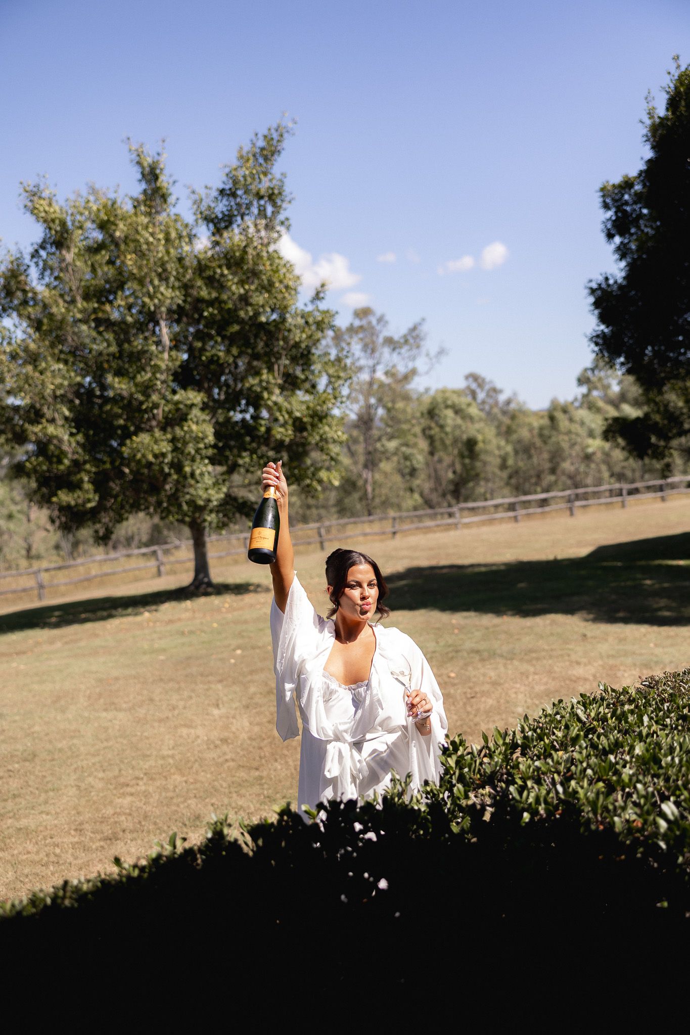 Bride in white robe raises Veuve Clicquot champagne bottle outdoors on sunny green lawn with trees.