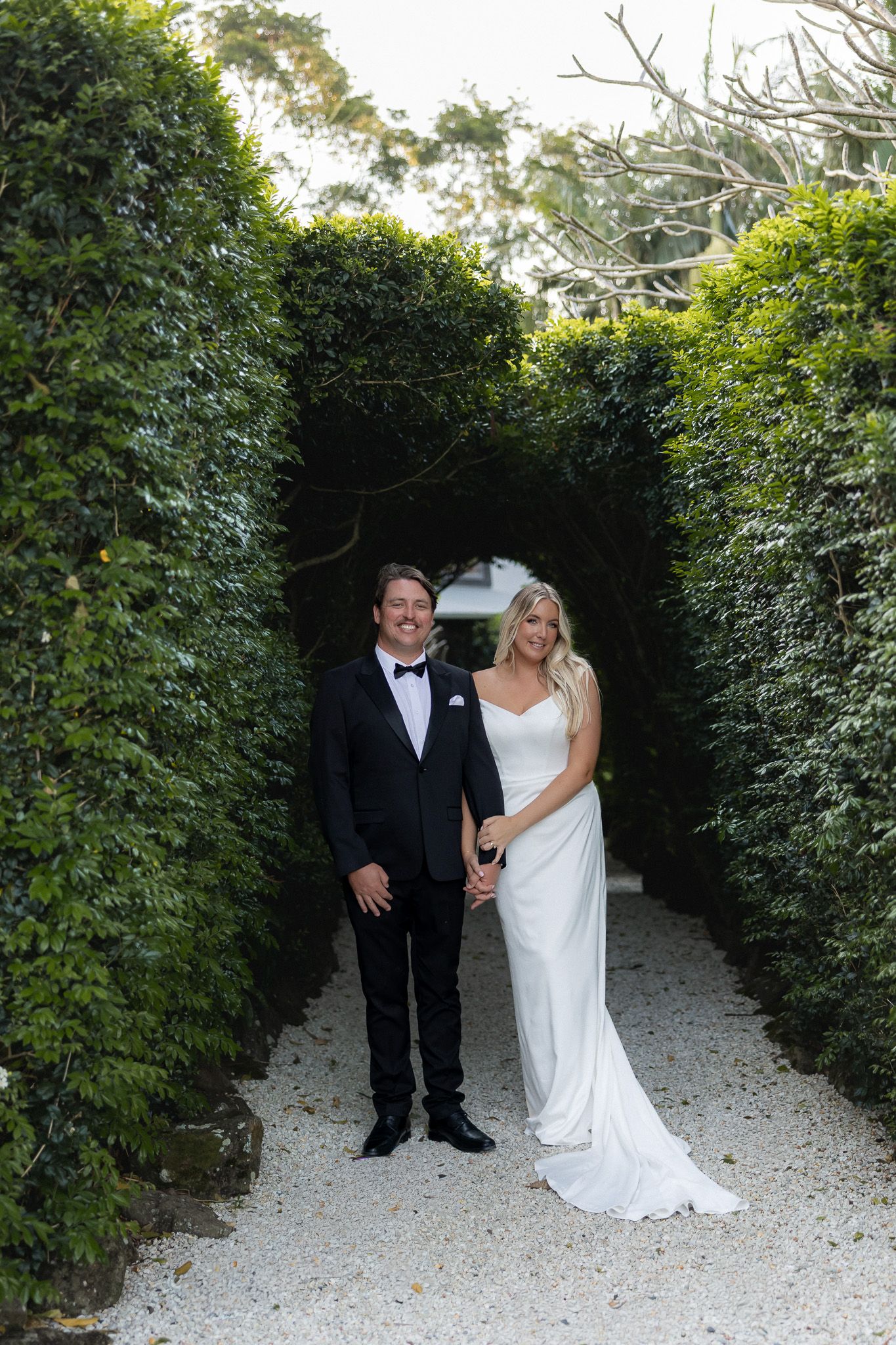 Bride and groom hold hands smiling in a romantic arched hedge walkway on their wedding day.